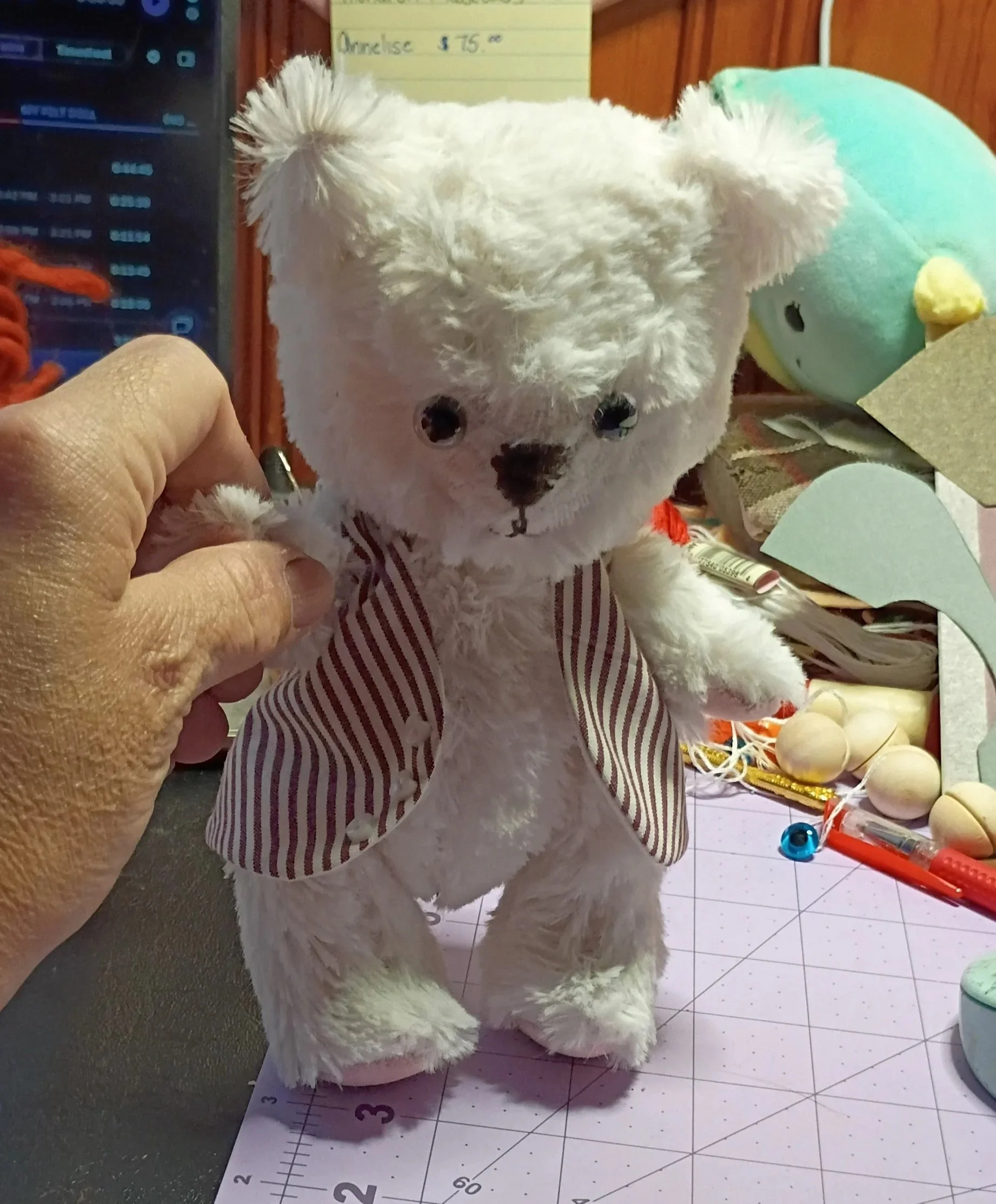 A white plush teddy bear with black eyes and a black nose, dressed in a red and white striped suit, is being held by a hand. The background includes a yellow paper with writing, a blue and green decorative ball, paper, and various objects on a checkered surface.