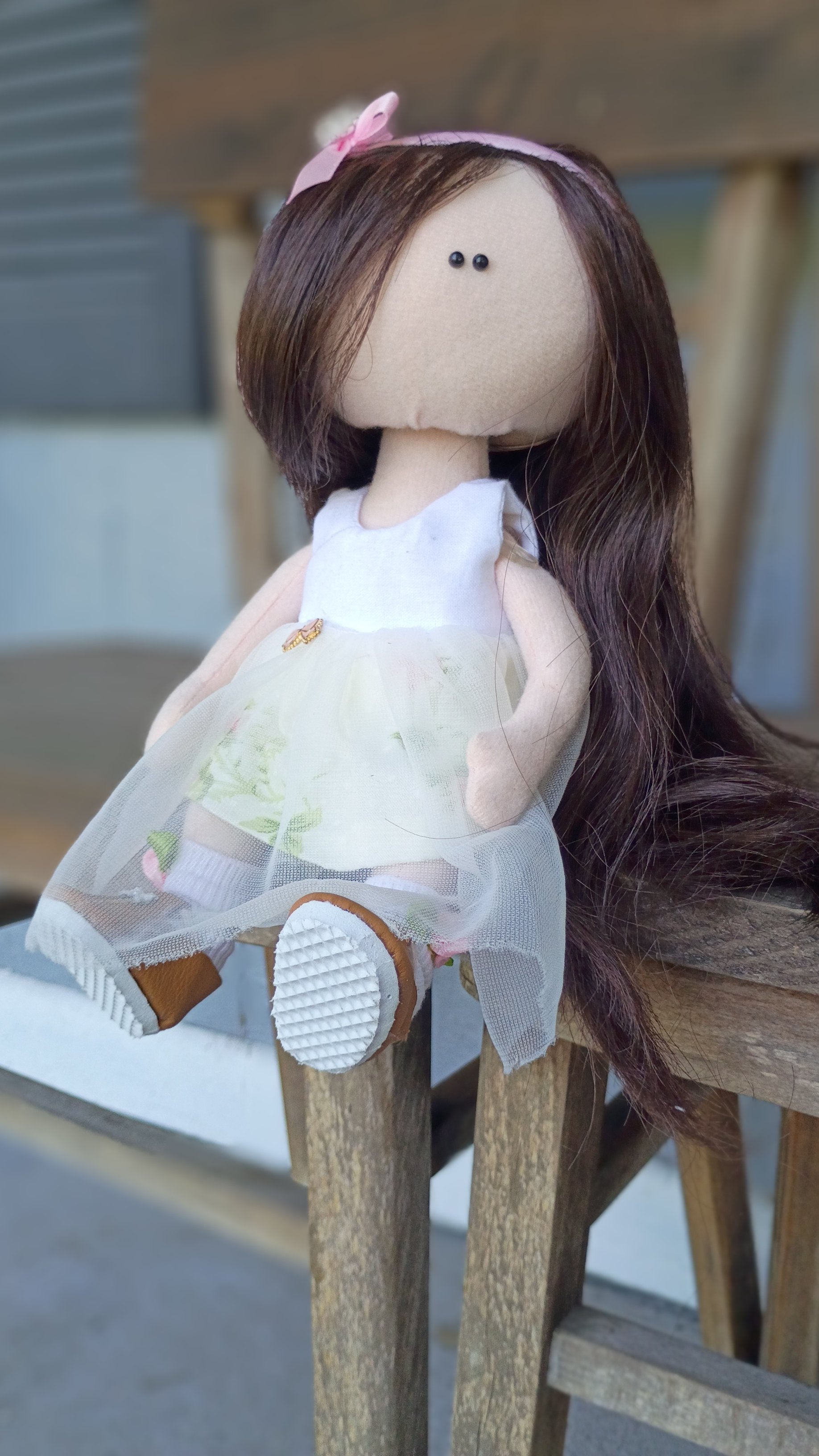A doll with a fabric face, brown hair, and black eyes, wearing a white dress with a floral design, sitting on a wooden bench with a pink bow headband and white sneakers.