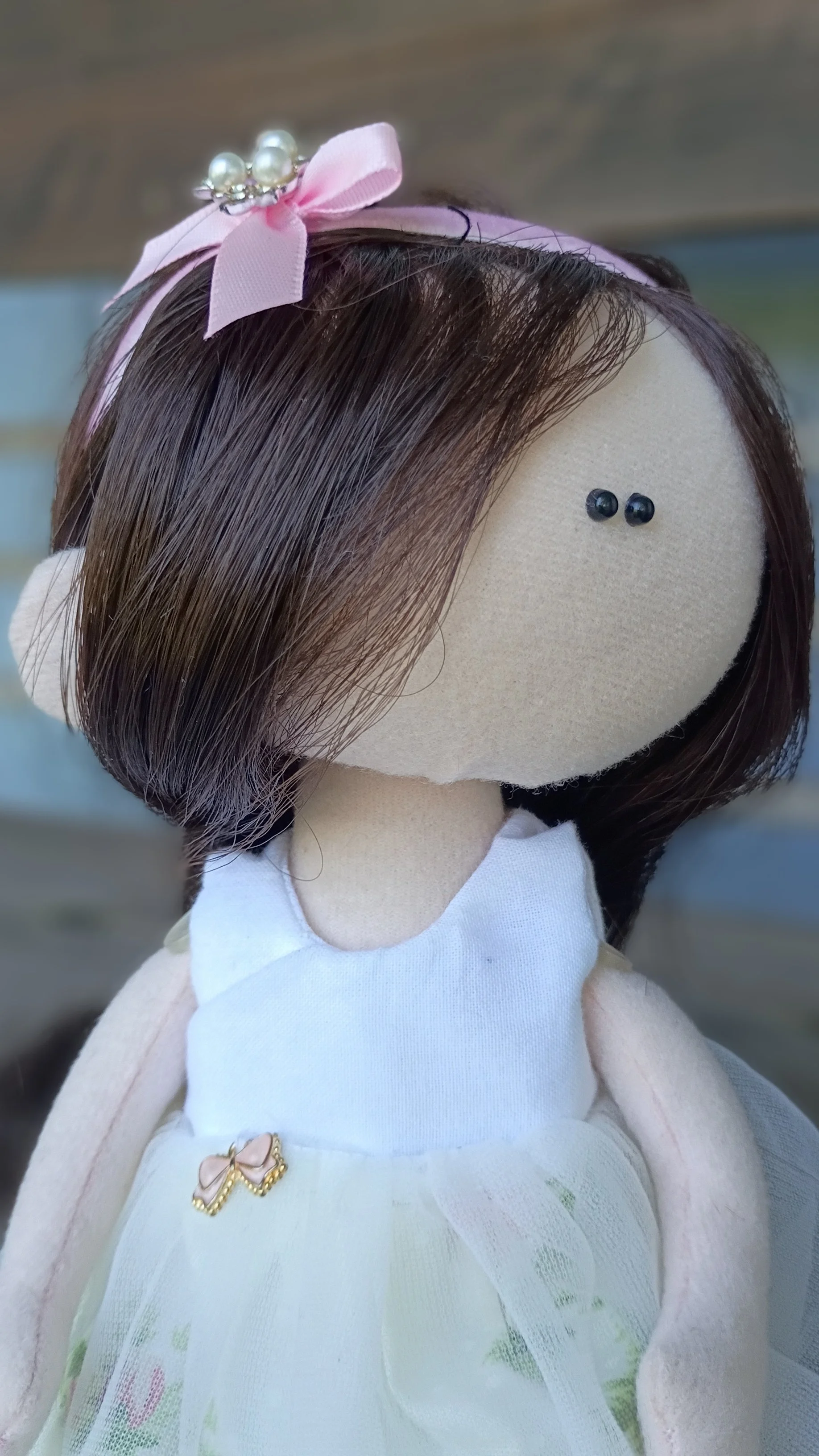 A handmade doll with brown hair, a pink ribbon with pearls on her head, black bead eyes, dressed in a white dress with a small bow, and a floral skirt, sitting against a blurred background.