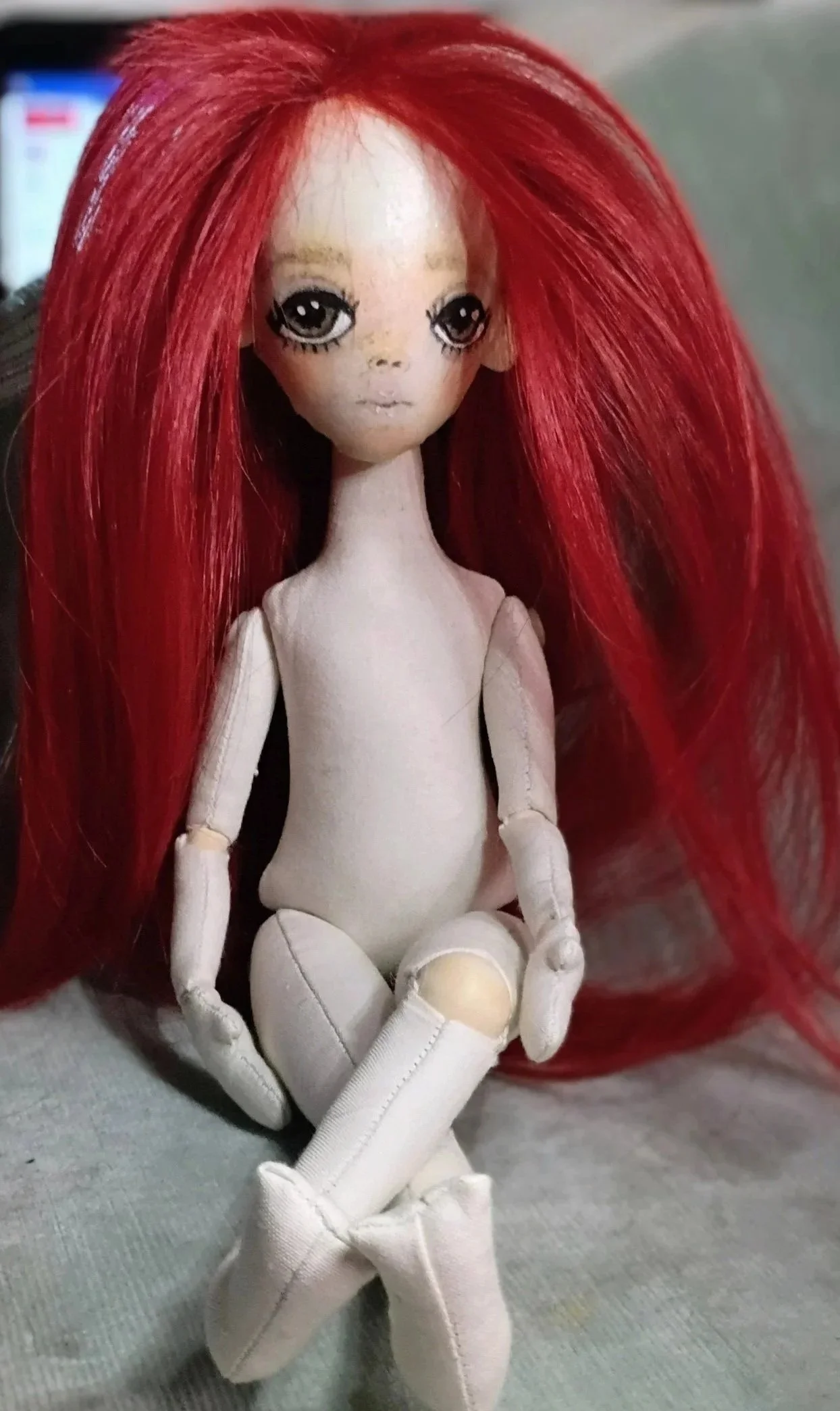 A handmade doll with large eyes and red hair, sitting with legs crossed and hands resting on knees.