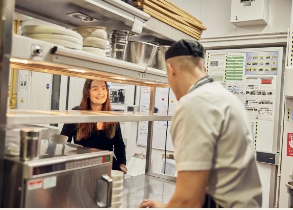 A young woman smiling at a chef behind a restaurant kitchen counter, with the woman in the background and the chef facing away.