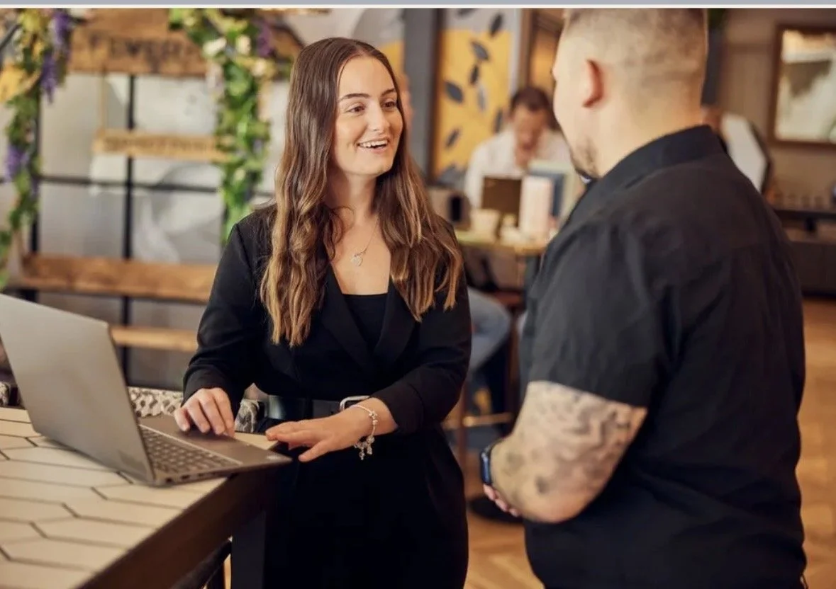 A woman with long brown hair, wearing a black dress with a bracelet and necklace, is smiling and talking to a man with short hair and a beard, dressed in black, in a cozy, decorated cafe.