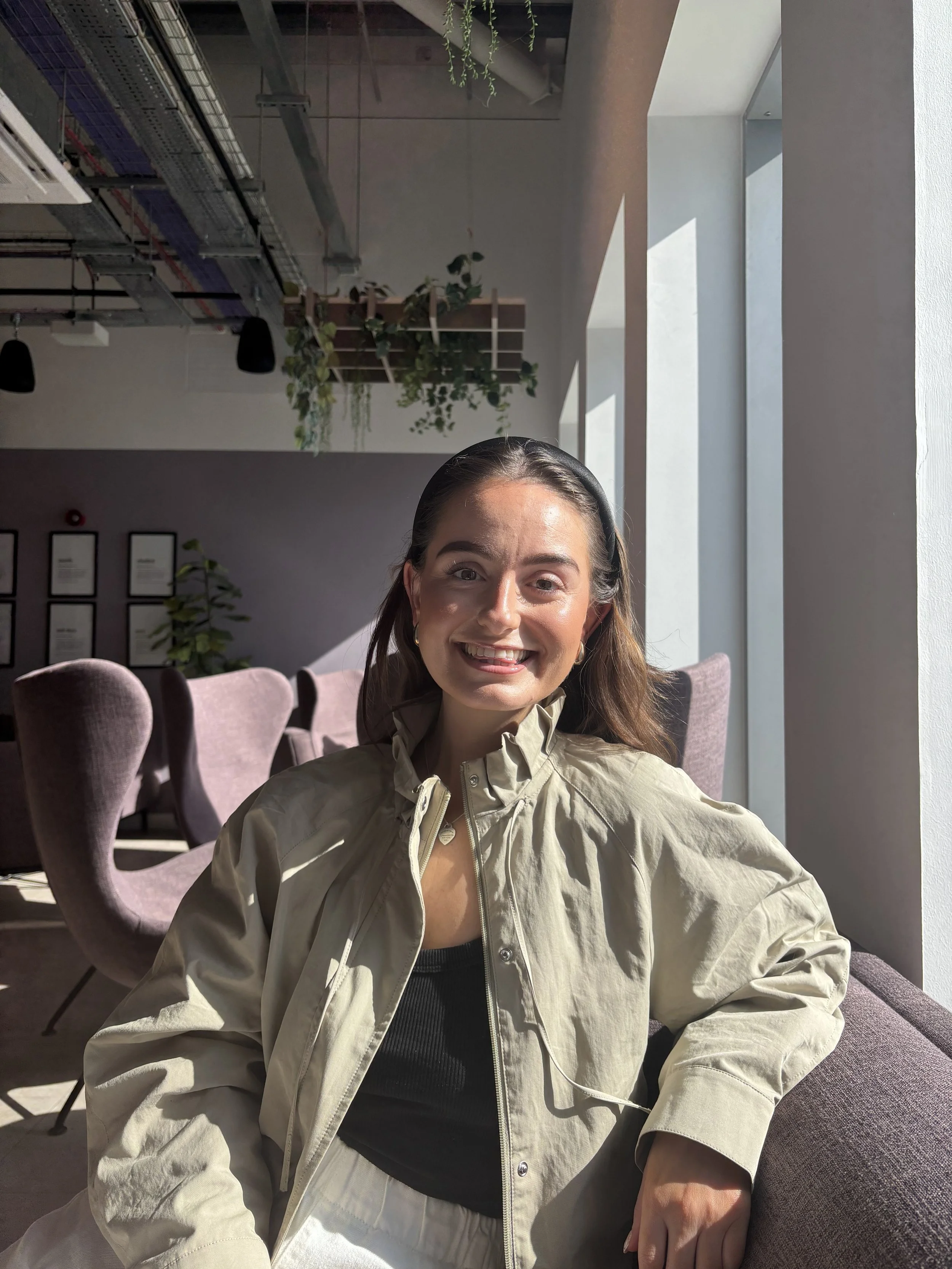 A woman with brown hair smiling, wearing a beige jacket and black top, seated in a modern room with purple chairs, large windows, and indoor plants, sunlight streaming in.