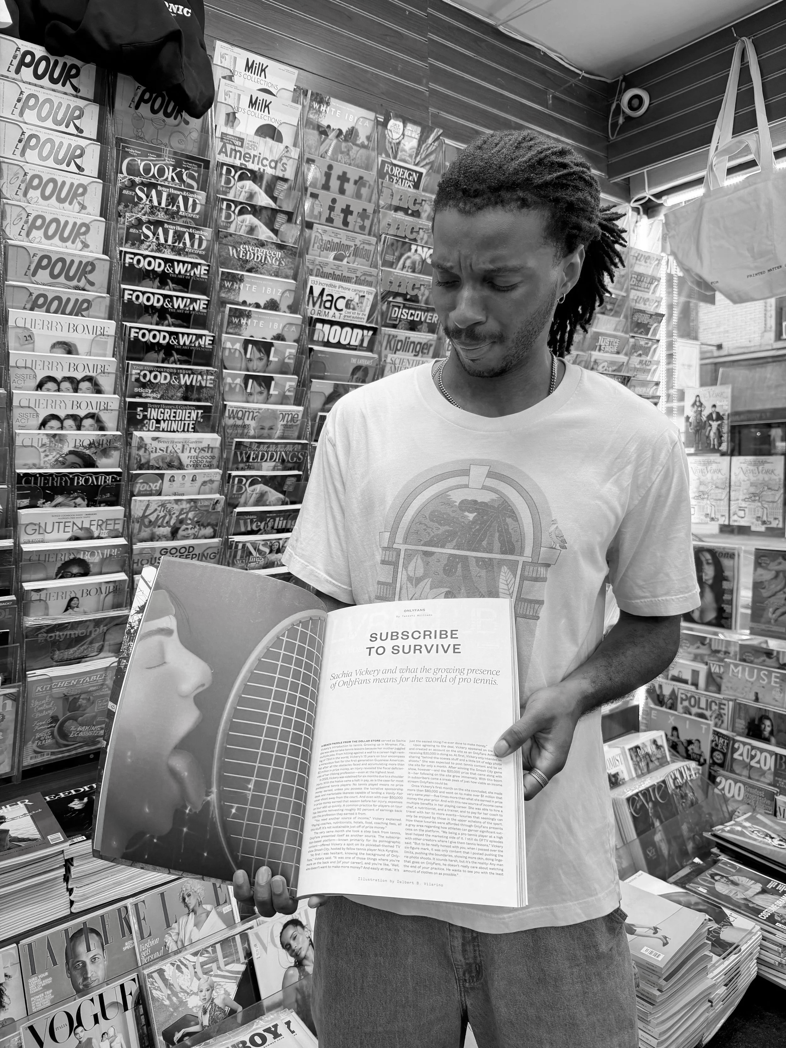 A man with dreadlocks holding open a magazine in a bookstore or newsstand, with magazine racks displaying various magazines and newspapers behind him.