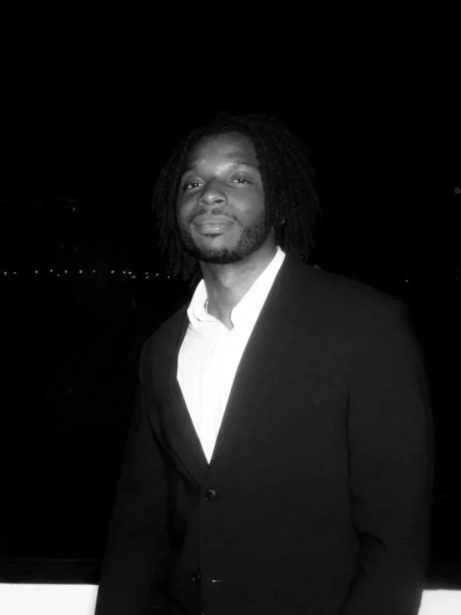 A man with shoulder-length dreadlocks wearing a white shirt and a dark blazer, standing outdoors at night.