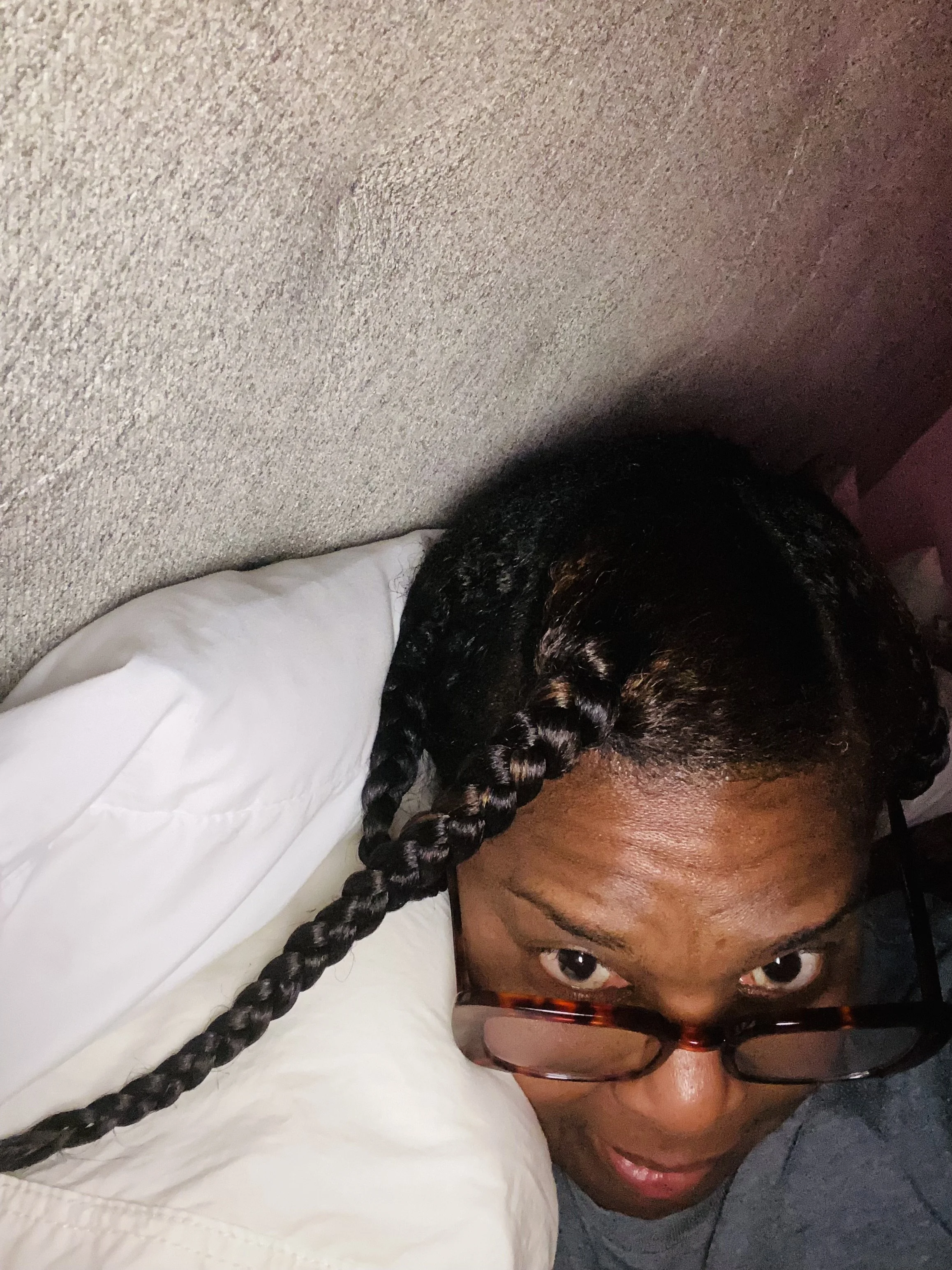 A woman with glasses and braided hair resting her head on a pillow, looking at the camera.