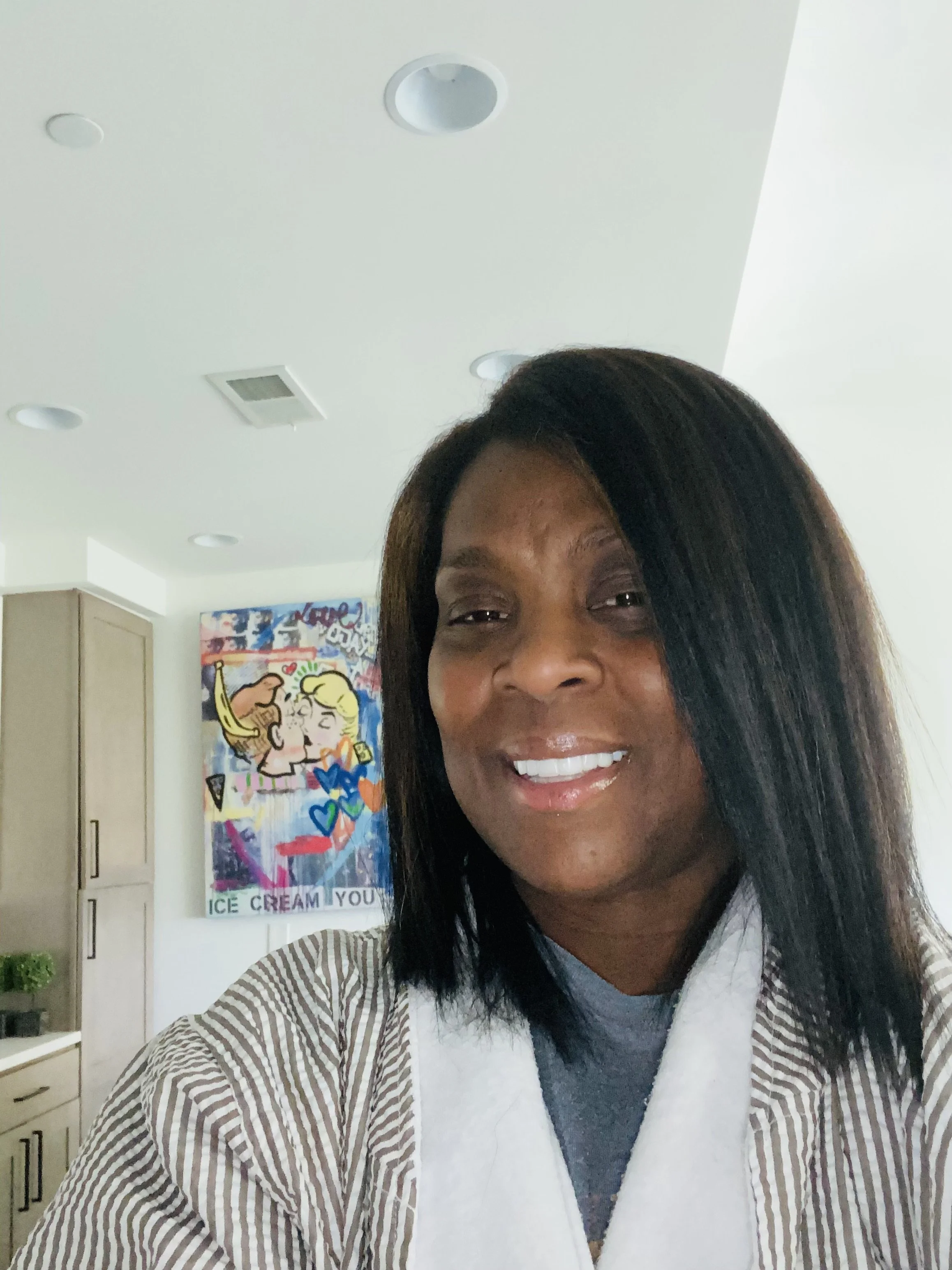 A woman with dark hair smiling for a selfie in a room with a colorful pop art style painting on the wall that says 'ICE CREAM YOU' and shows two cartoon characters of a man and woman kissing.