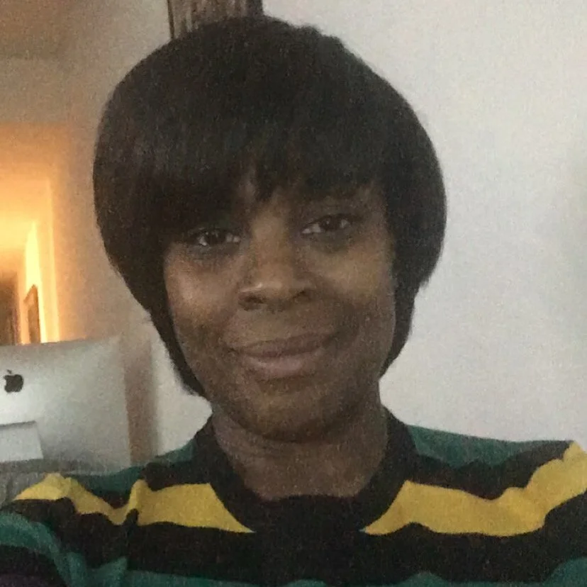 A woman with short black hair smiling, wearing a black, green, and yellow striped shirt indoors near a white wall.