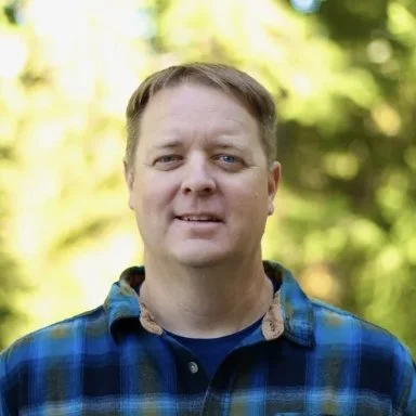 Portrait of a man outdoors with blurred trees and sunlight in the background.