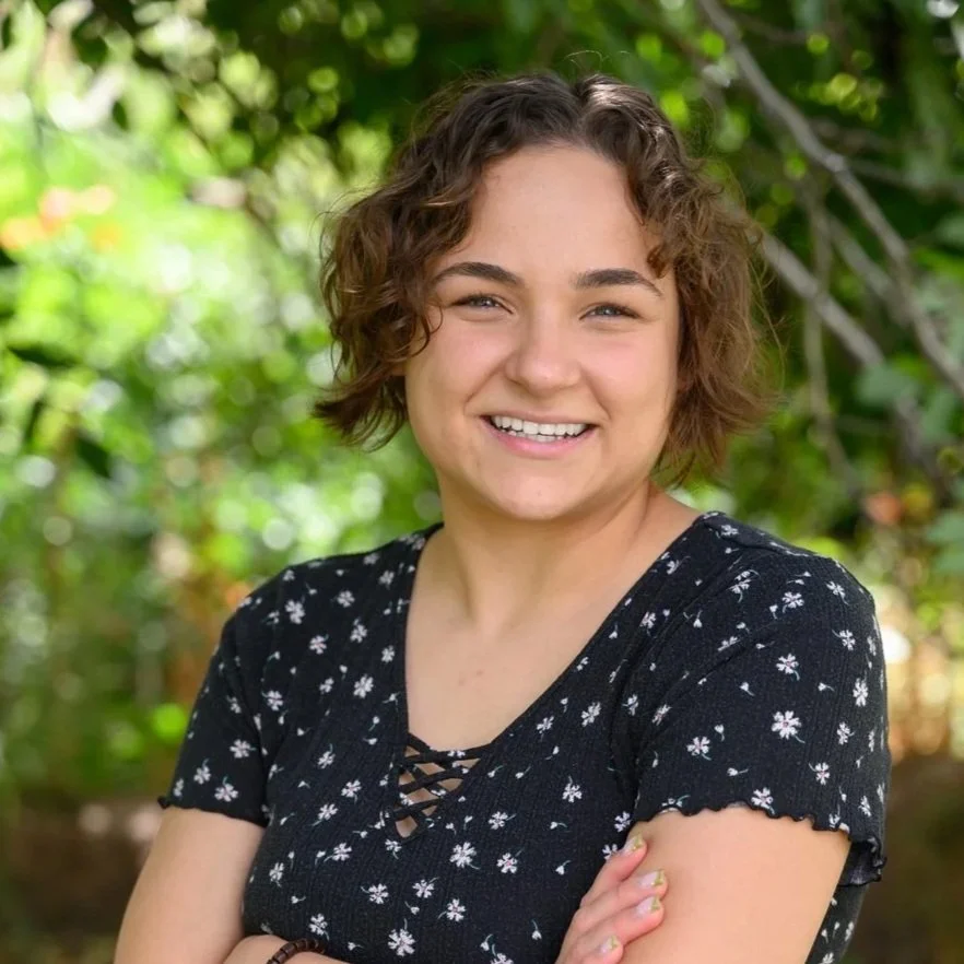 A young woman with curly brown hair smiling outdoors with green foliage in the background.