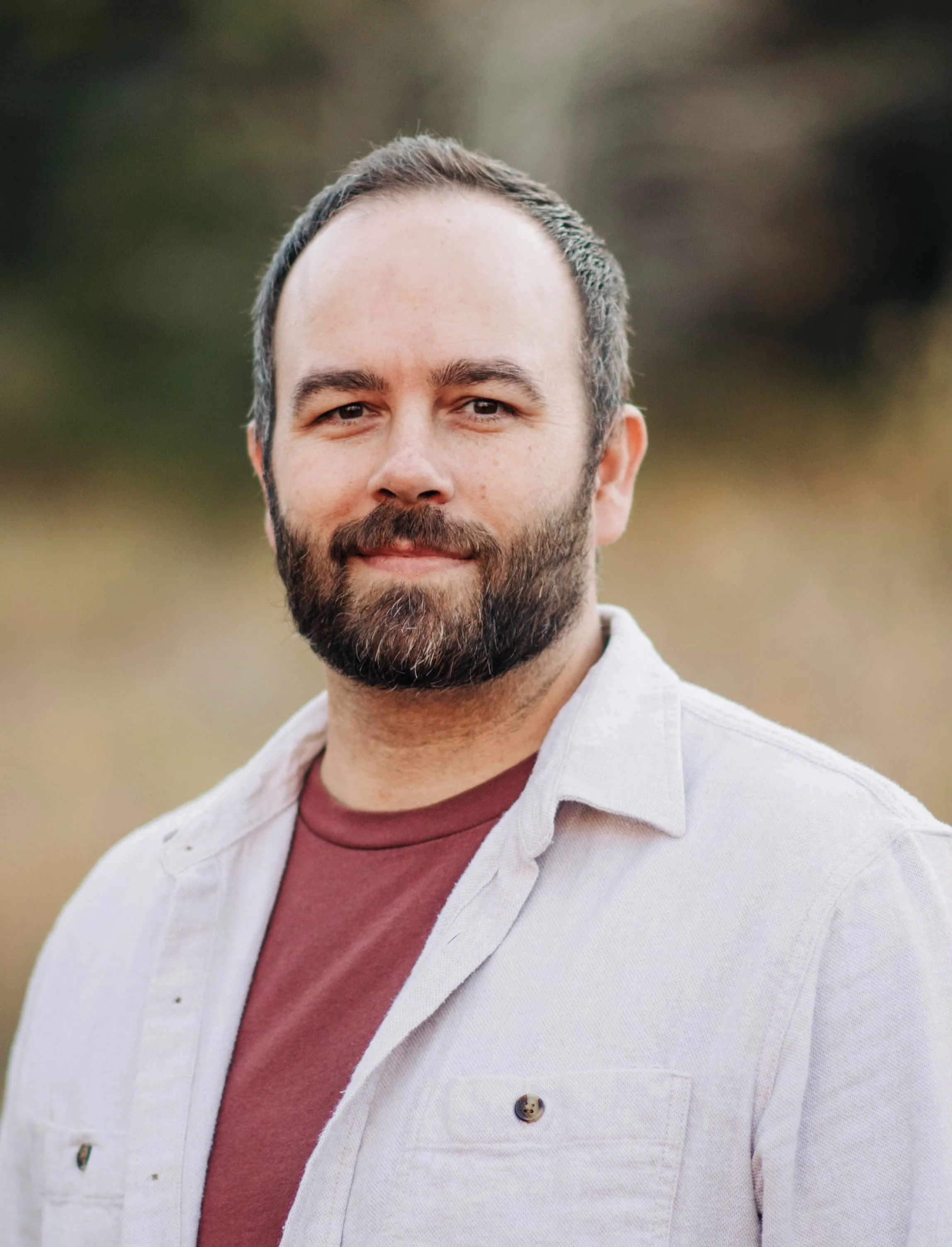 Close-up of a man with a beard and short dark hair, wearing a beige jacket over a maroon shirt, standing outdoors against a blurred background of greenery.