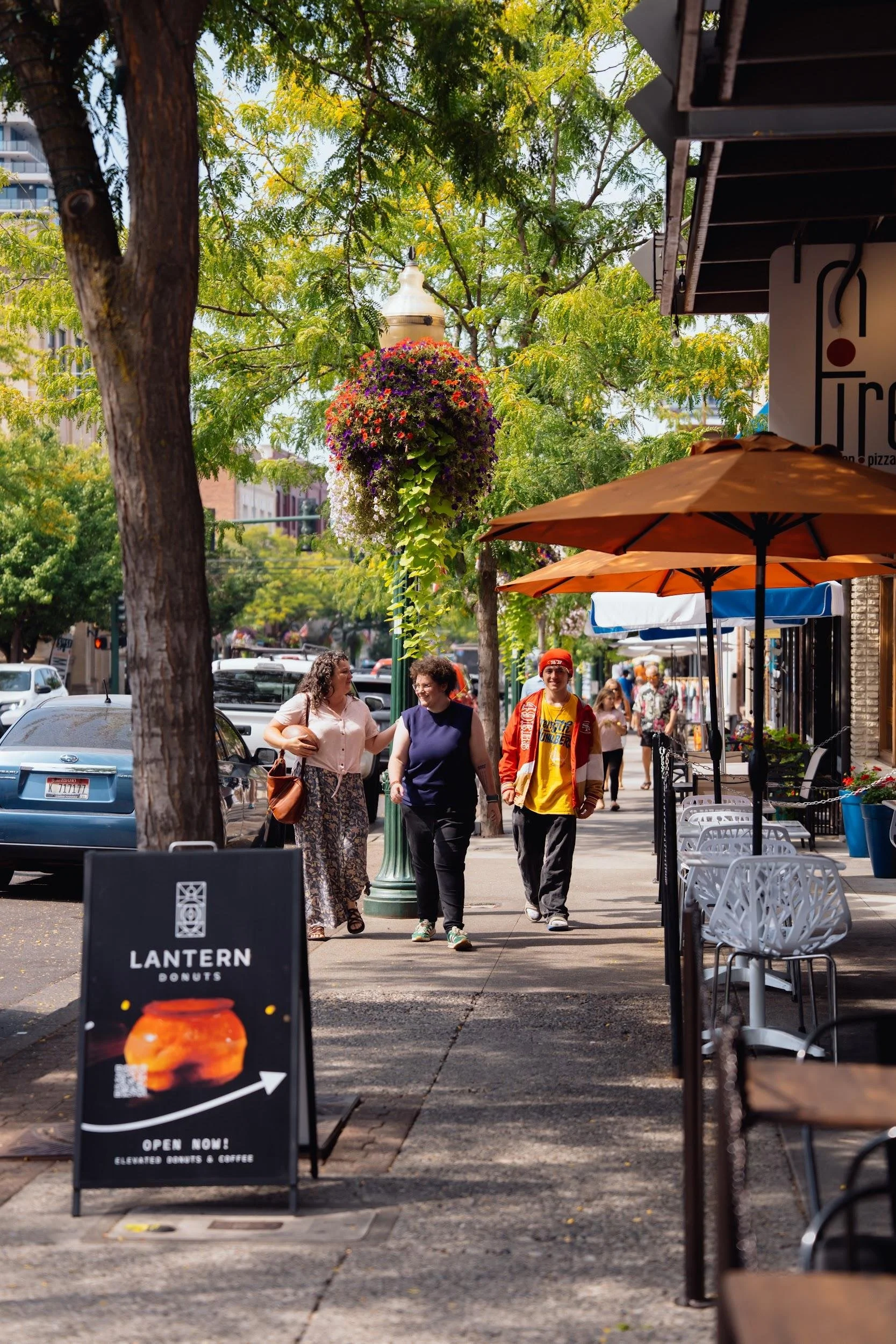 City sidewalk with people walking, outdoor seating with orange umbrellas, a flower basket hanging from a lamp post, and a sign for Lantern Donuts.