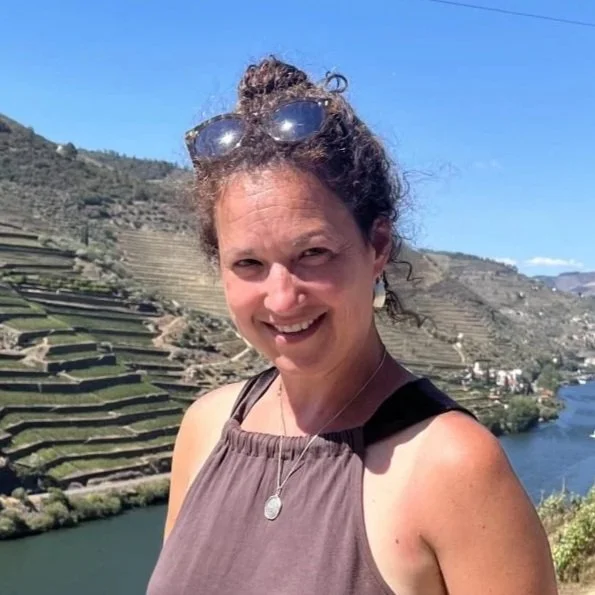 A woman with curly hair, wearing sunglasses on her head, smiling outdoors in front of terraced hills and a river.