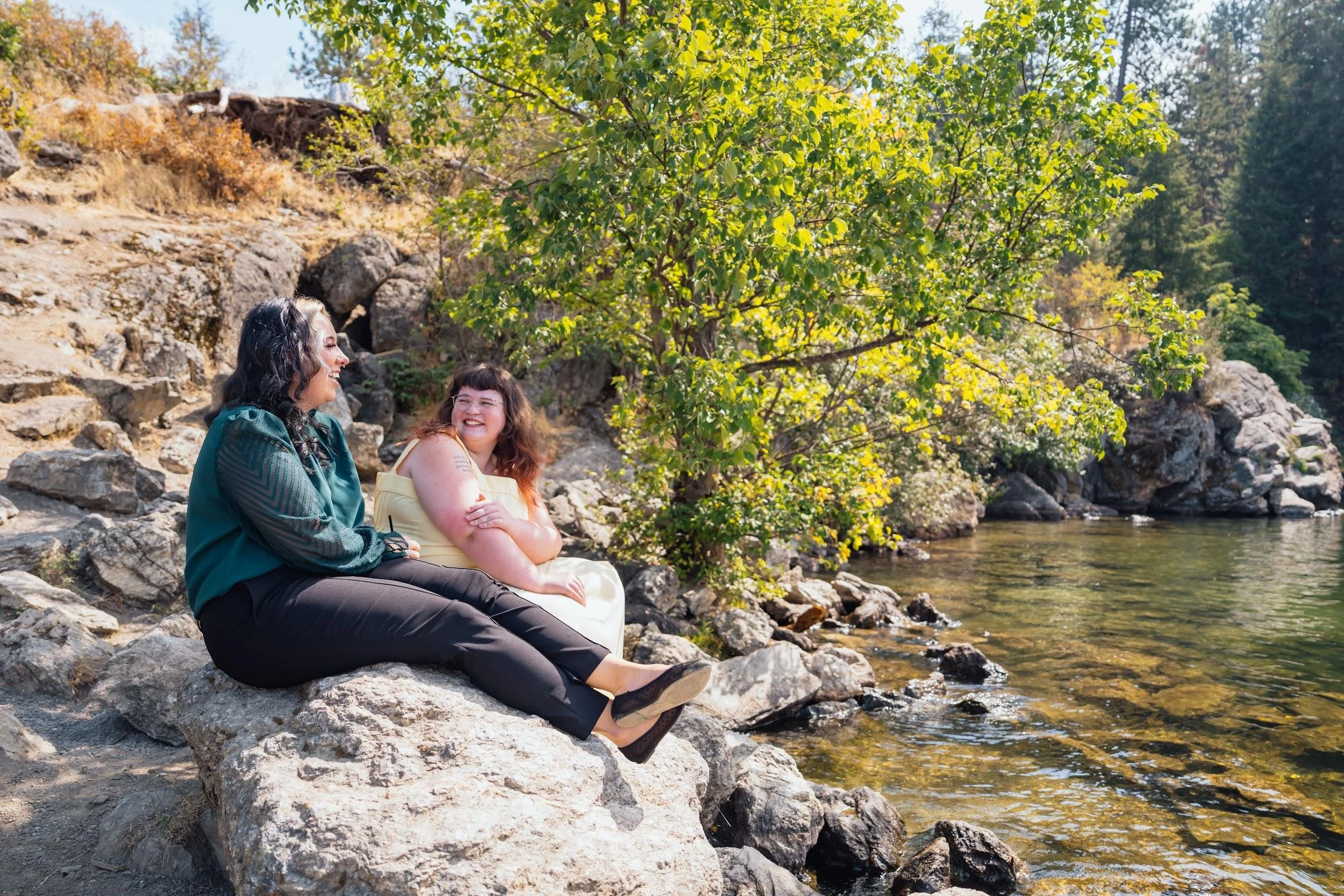 Two women sitting on rocks by a river, smiling and enjoying a sunny day surrounded by trees and nature.