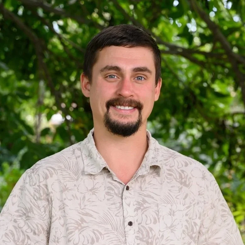 A smiling man with dark hair, a beard, and mustache, wearing a beige short-sleeved, floral-patterned shirt, standing outdoors with green foliage in the background.