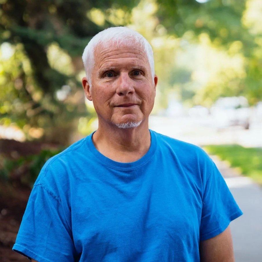 A man with short white hair and a beard wearing a blue t-shirt standing outdoors with trees and sunlight in the background.