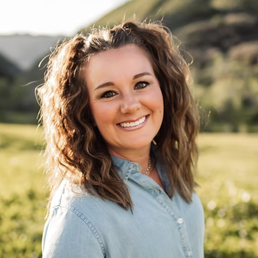 A smiling woman with curly brown hair wearing a light blue denim shirt outdoors with green hills in the background.