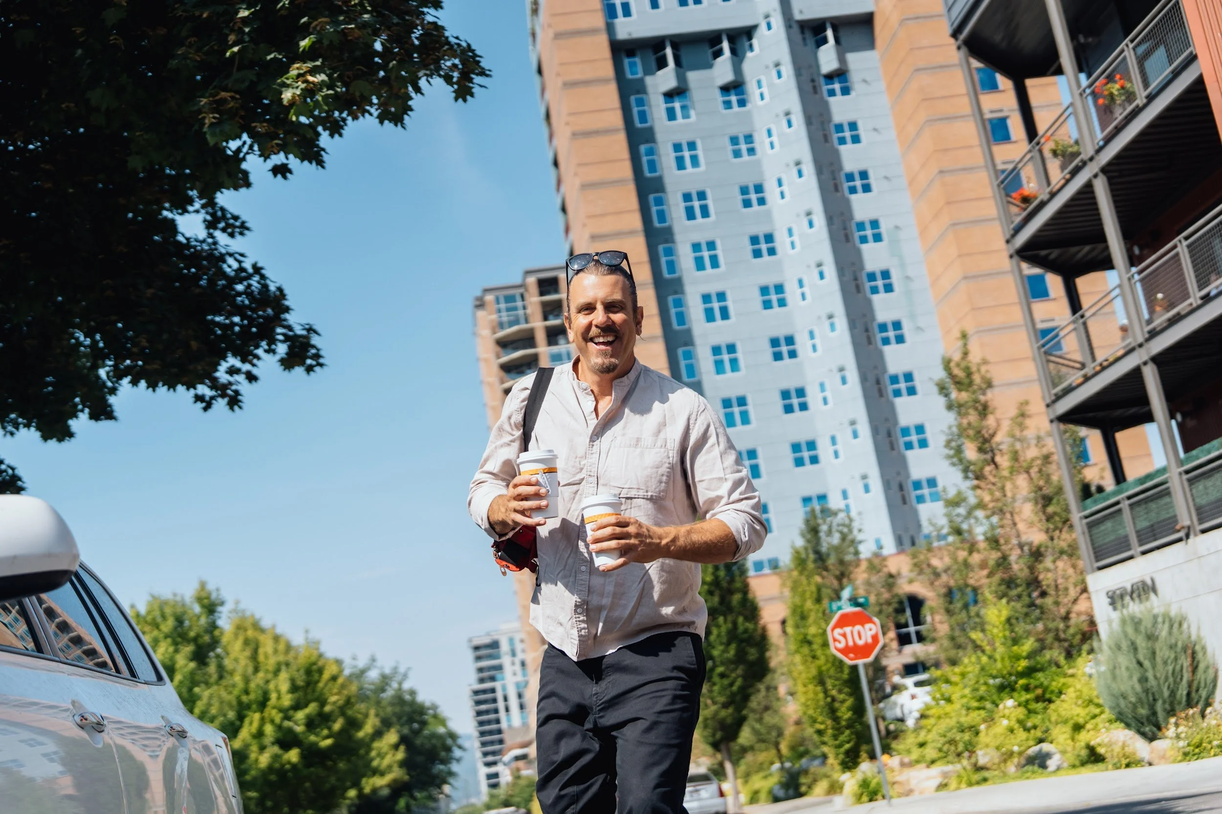 Smiling man carrying two coffee cups on a sunny street in downtown Coeur d'Alene