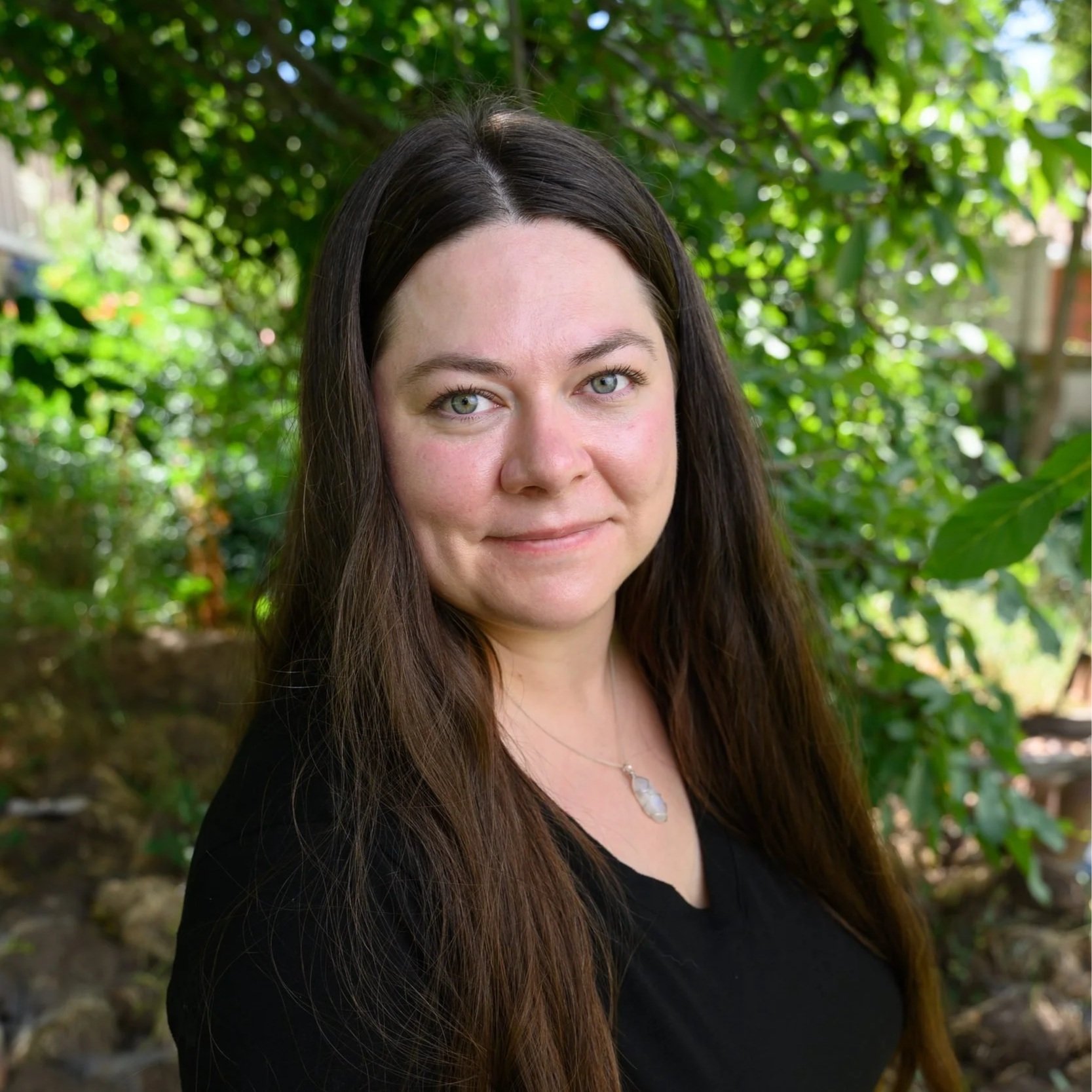 Close-up of a woman with long brown hair and blue eyes outdoors with green foliage in the background.