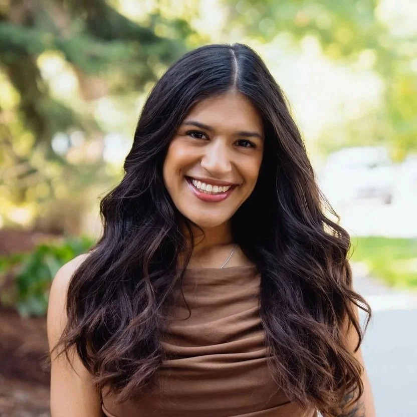 A woman with long wavy black hair smiling outdoors with trees and greenery in the background.