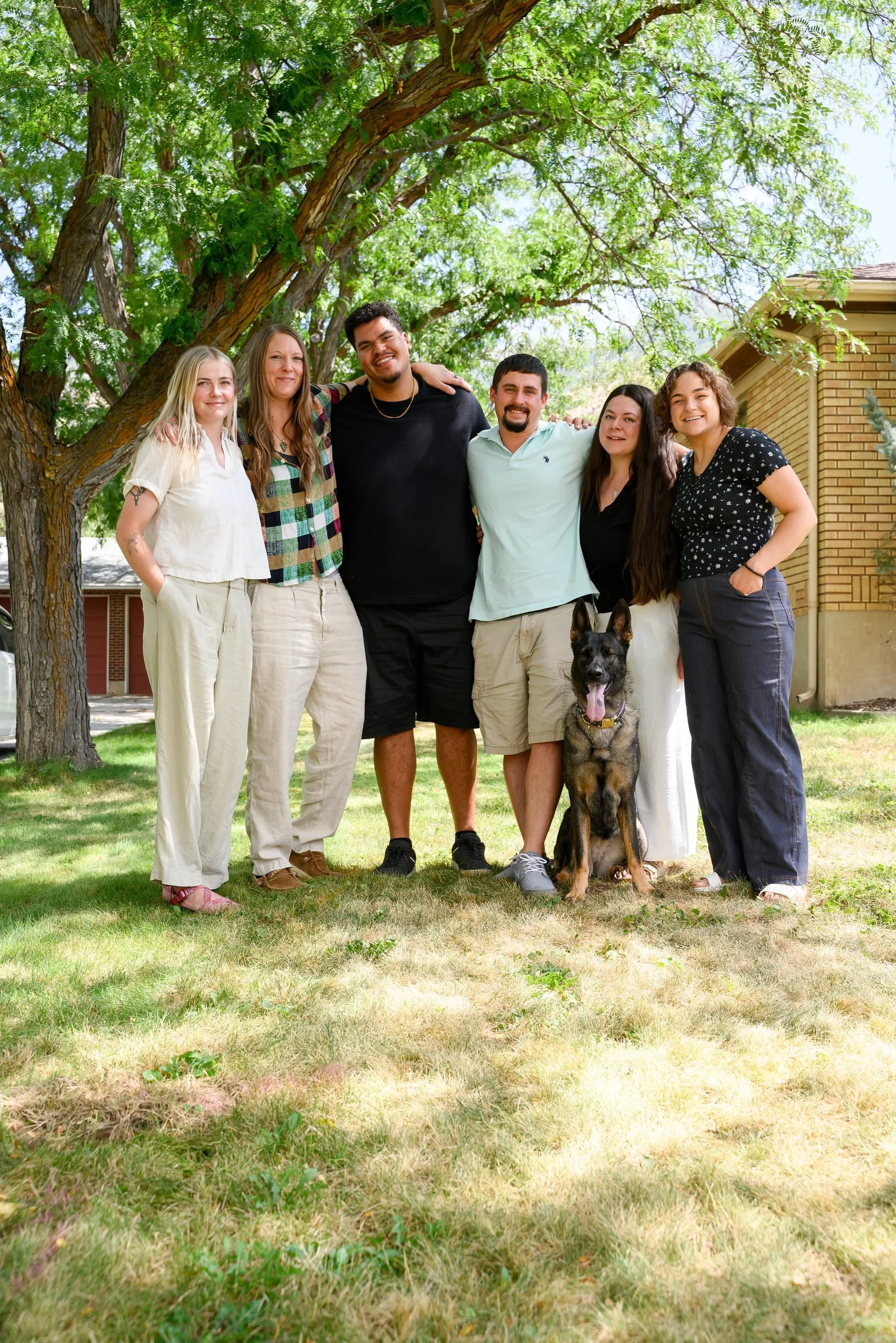 Group of six diverse people, four women and two men, standing outdoors on grass under a large leafy tree with a dog sitting in front of them, near a brick house, smiling for a photo on a sunny day.