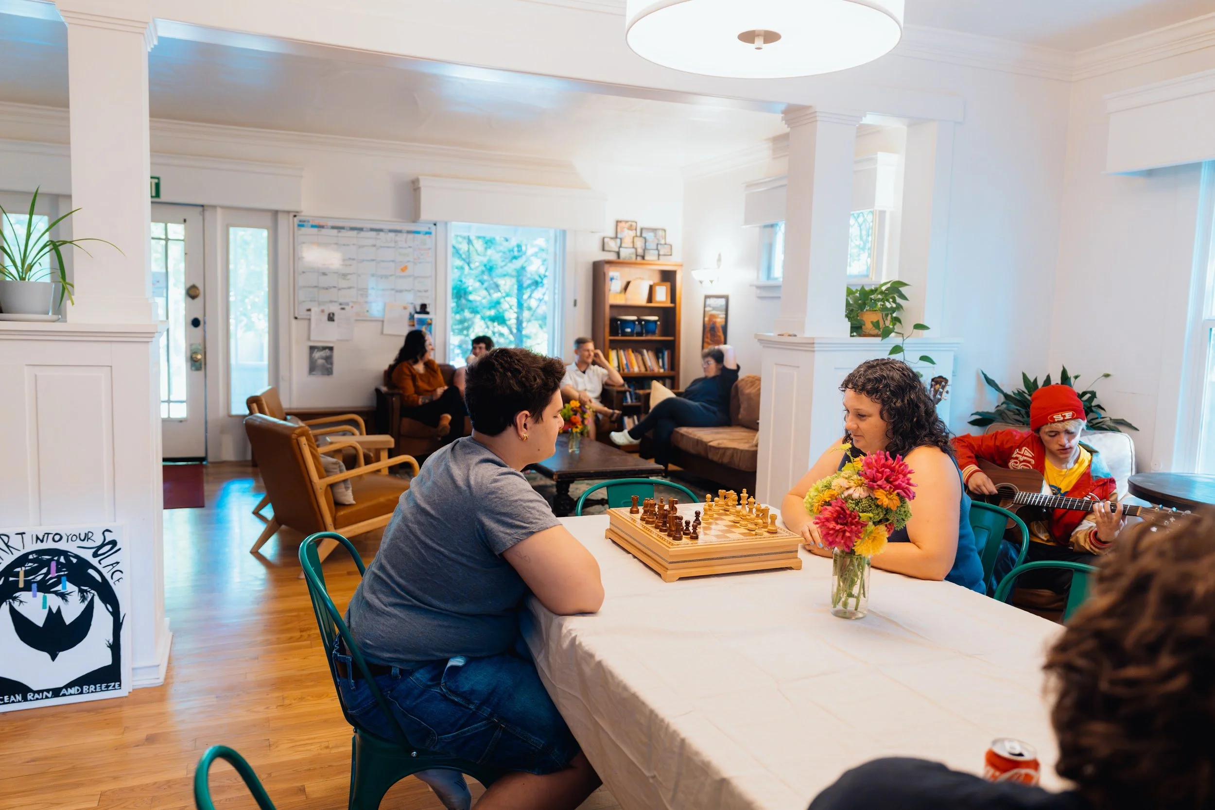 Two young adults playing chess at a dining table while others talk in the living room