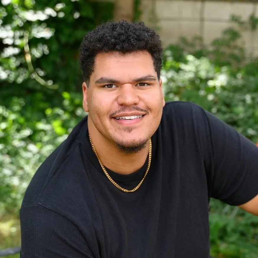 A young man with dark curly hair, a beard, and mustache smiling outdoors in front of green foliage, wearing a black shirt and a gold chain necklace.
