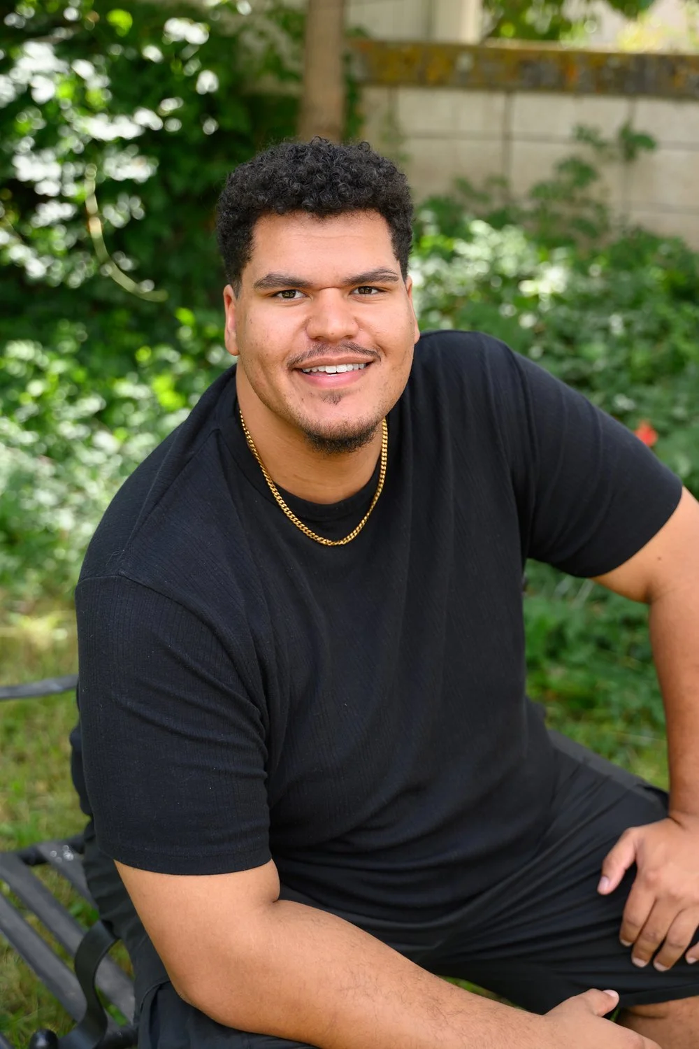 A smiling man with curly hair, wearing a black t-shirt and a gold chain, sitting outdoors in a lush garden.