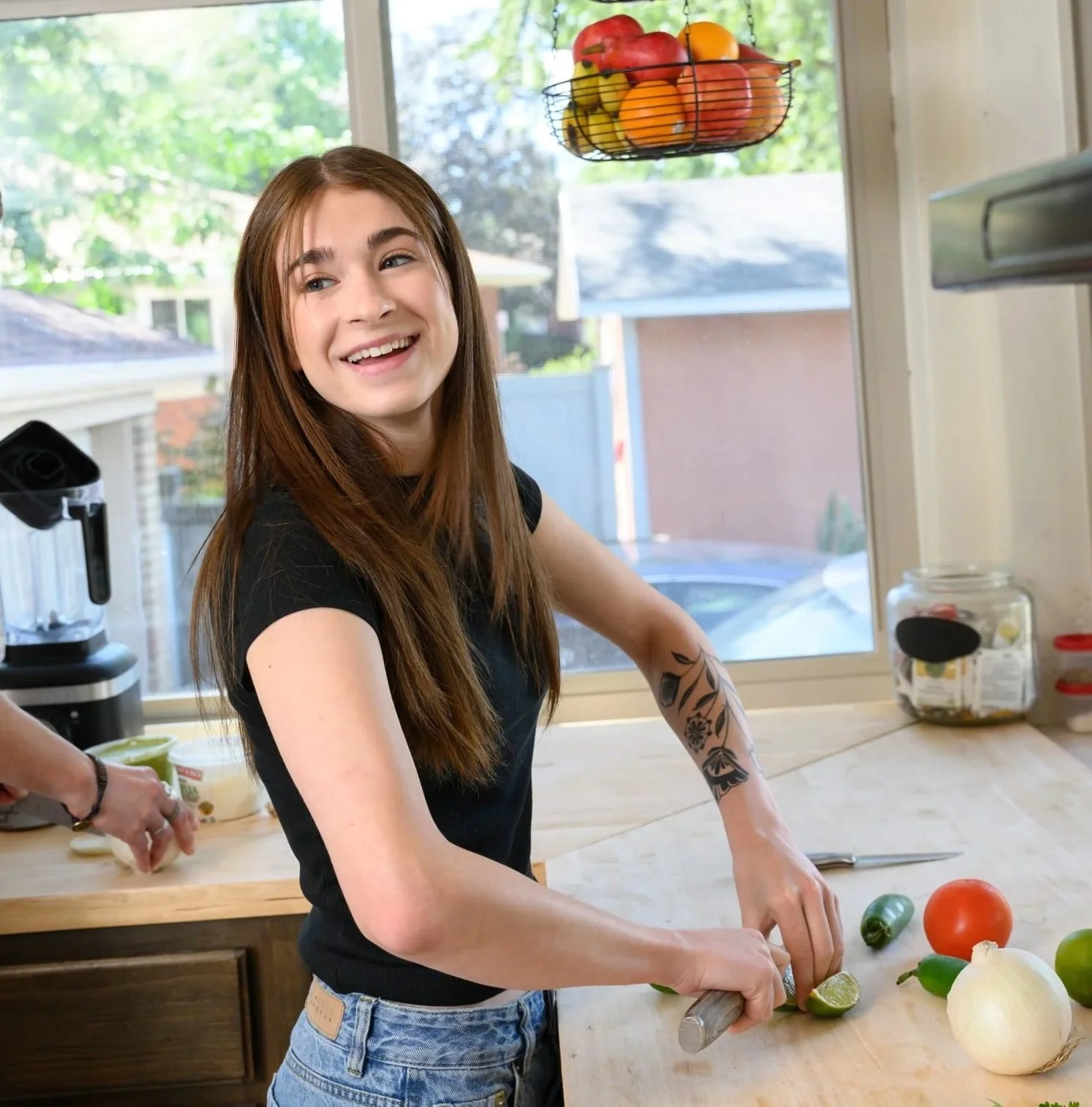 A young woman with long brown hair and a tattoo on her arm, smiling while preparing vegetables in a kitchen. She is cutting a lime on a wooden countertop, with tomatoes, onions, and peppers around her. There is a window behind her showing trees and houses outside.