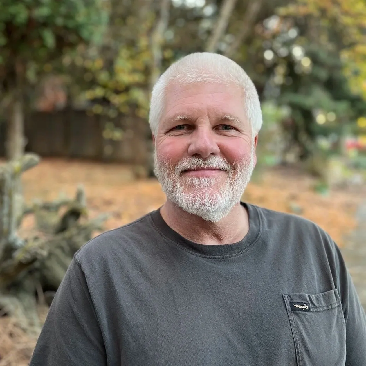 A middle-aged man with white hair and a beard smiling outdoors during fall, wearing a dark grey Wrangler t-shirt, with trees and fallen leaves in the background.