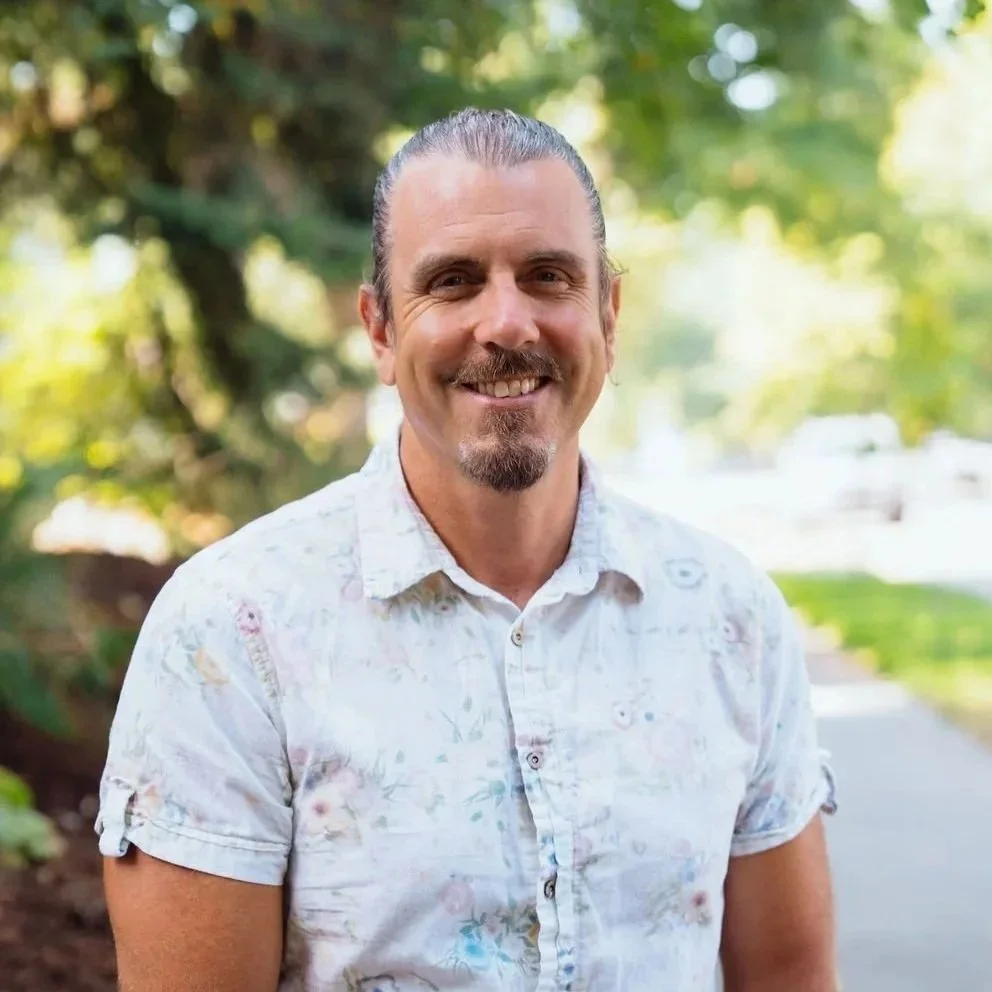 A man with dark blond hair slicked back, a mullet, and a goatee, smiling outdoors with trees and greenery in the background, wearing a light-colored, floral short-sleeved shirt.