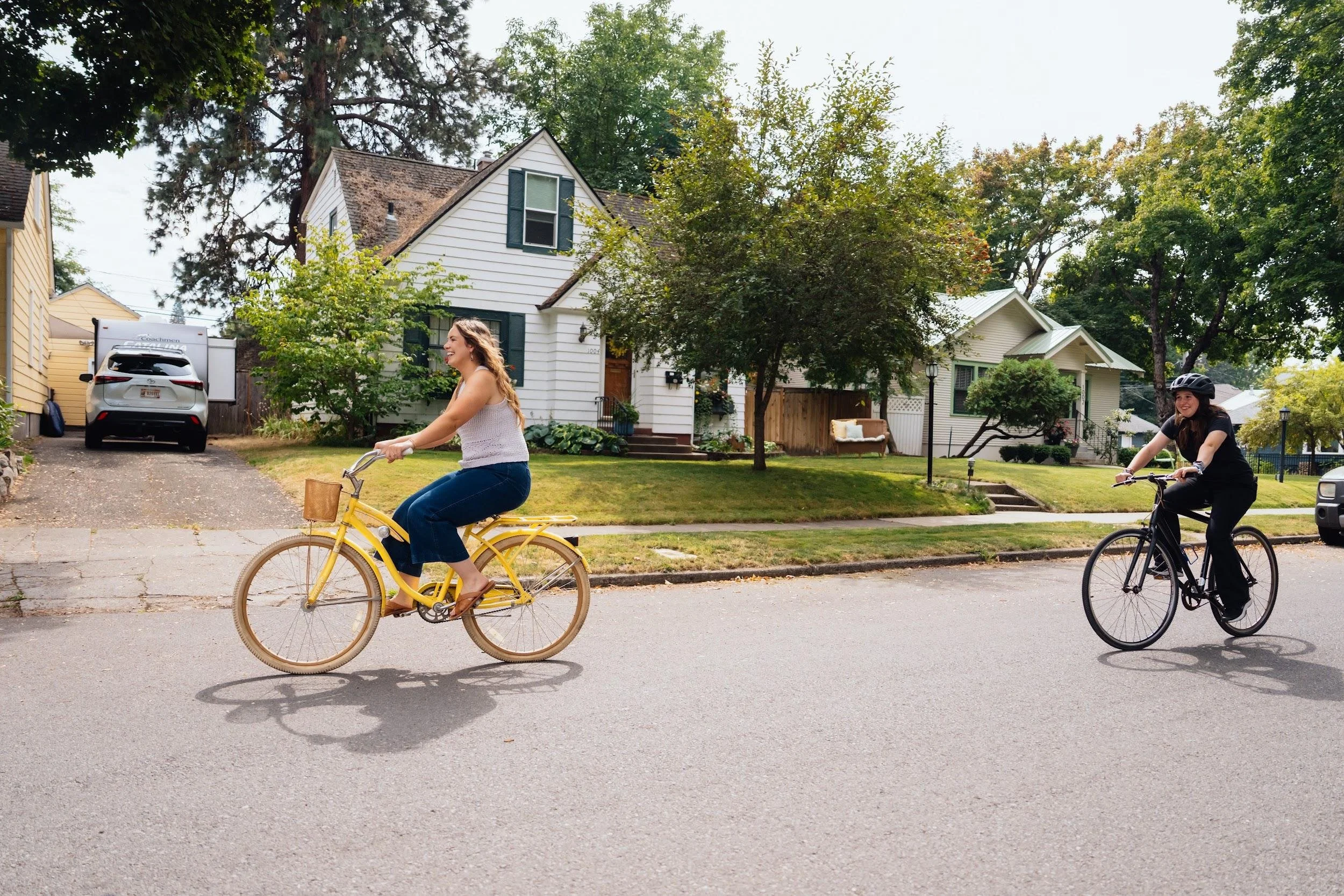 Young woman riding a yellow bicycle past a white house on a tree-lined street