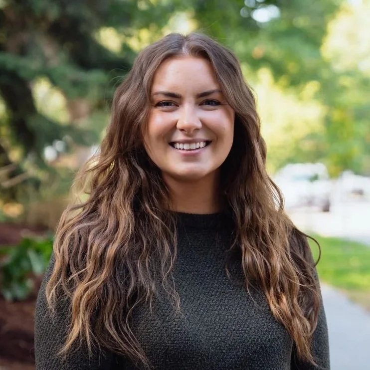 A smiling woman with long, wavy brown hair standing outdoors with trees and a blurred background.