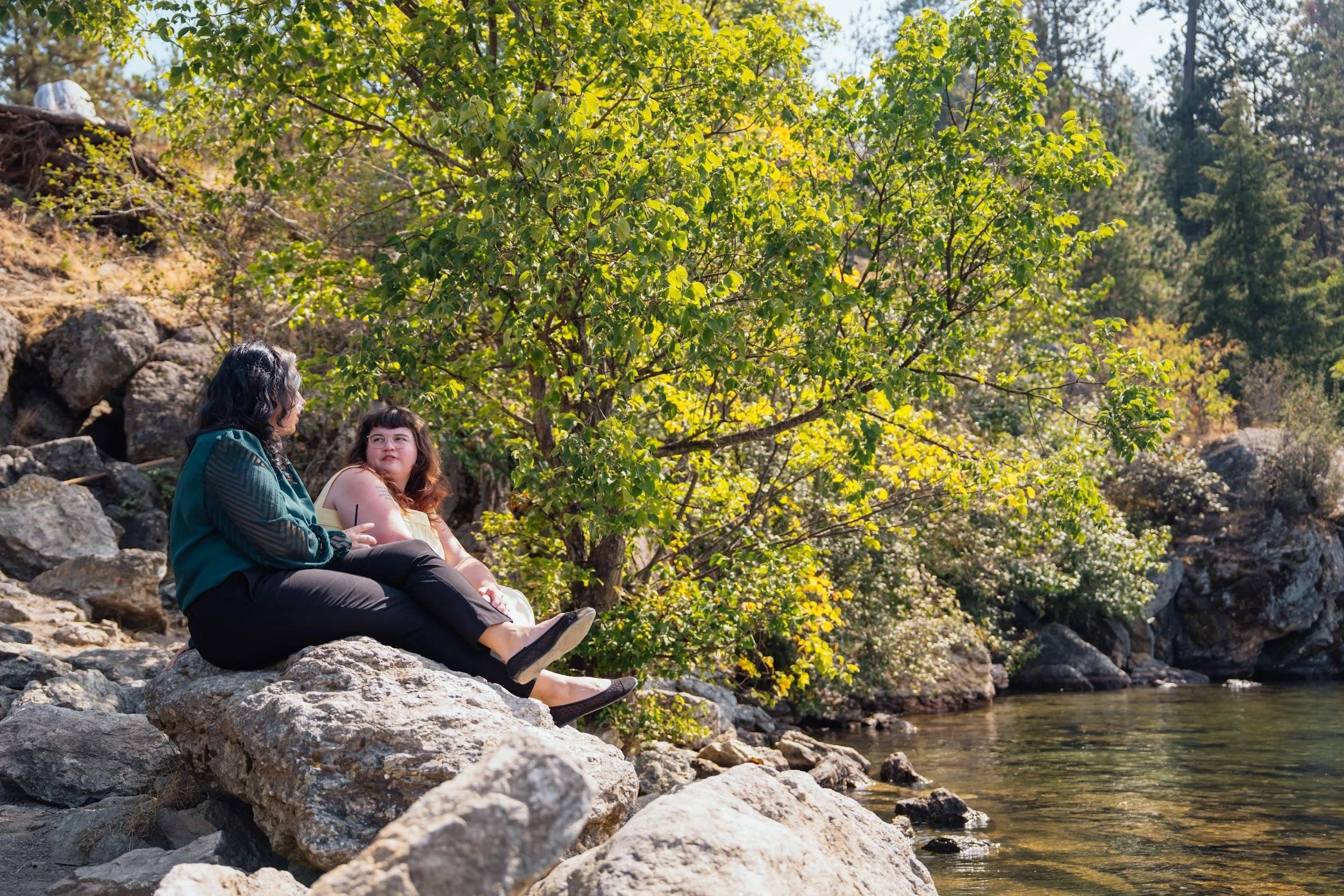 Two young women sitting on lakeshore rocks talking beneath green trees