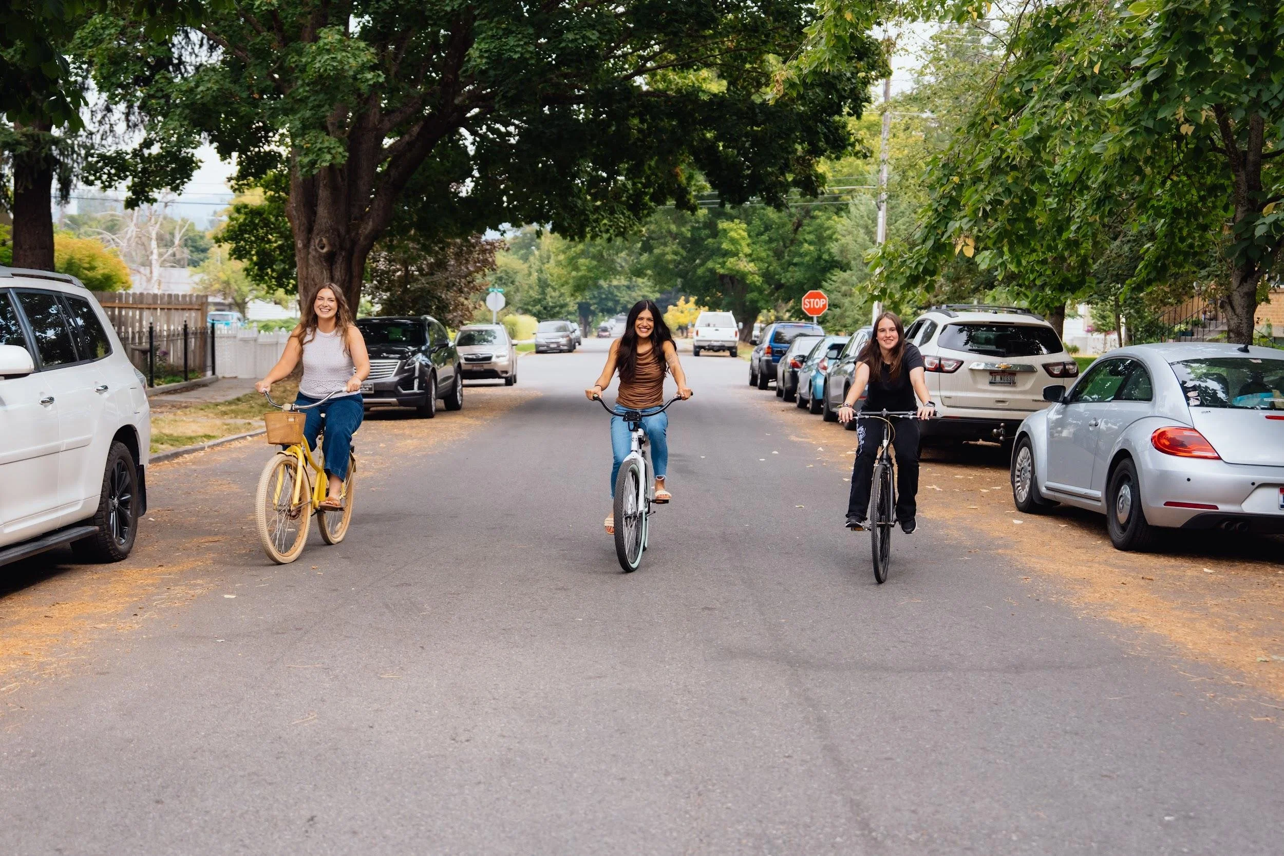 Three women riding bicycles down a suburban street lined with parked cars and green trees.