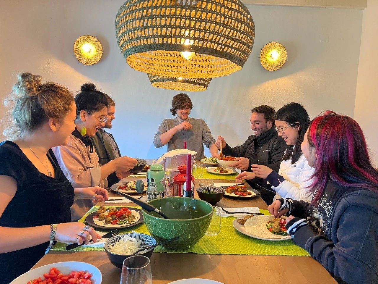 Group of people enjoying a meal together at a dinner table with various dishes and drinks, indoors with woven light fixtures on the ceiling.
