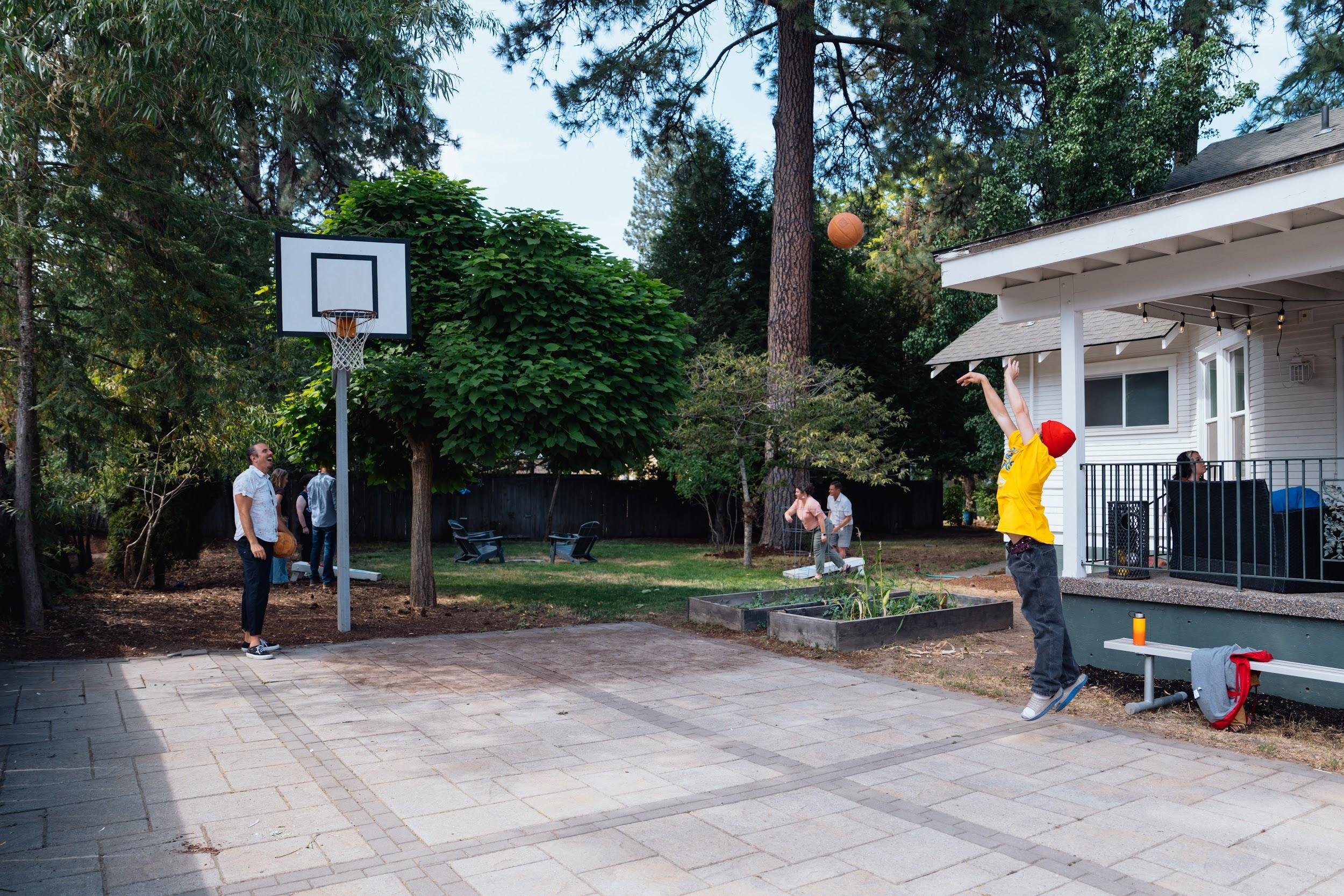 People playing basketball outdoors in a backyard, some people are watching, surrounded by trees and a house with a porch decorated with string lights.