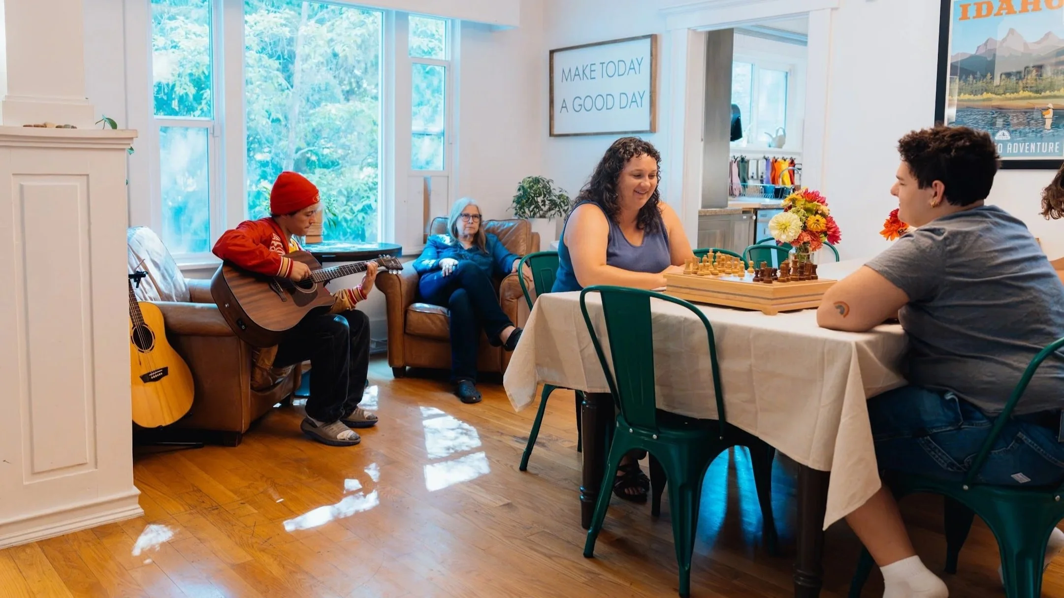 People playing chess at a dining table and listening to a musician playing guitar in a cozy, well-lit room.