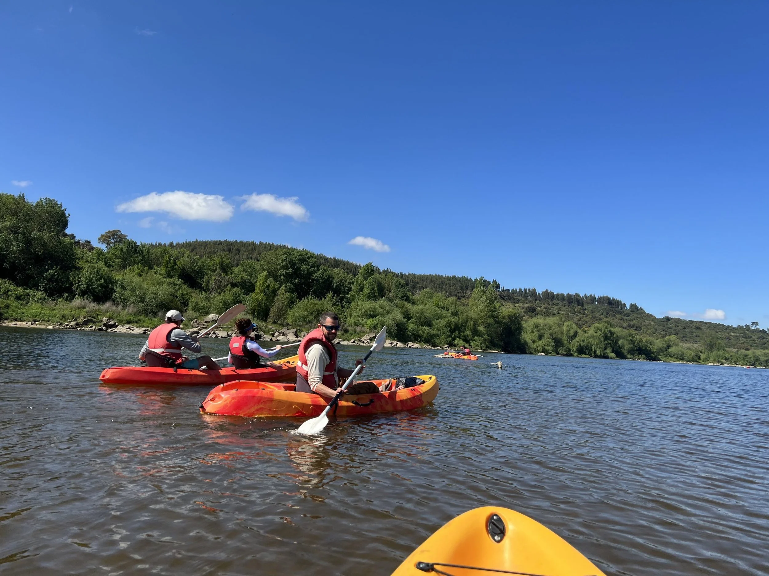 People kayaking on a calm river surrounded by green trees and hills under a blue sky with white clouds.
