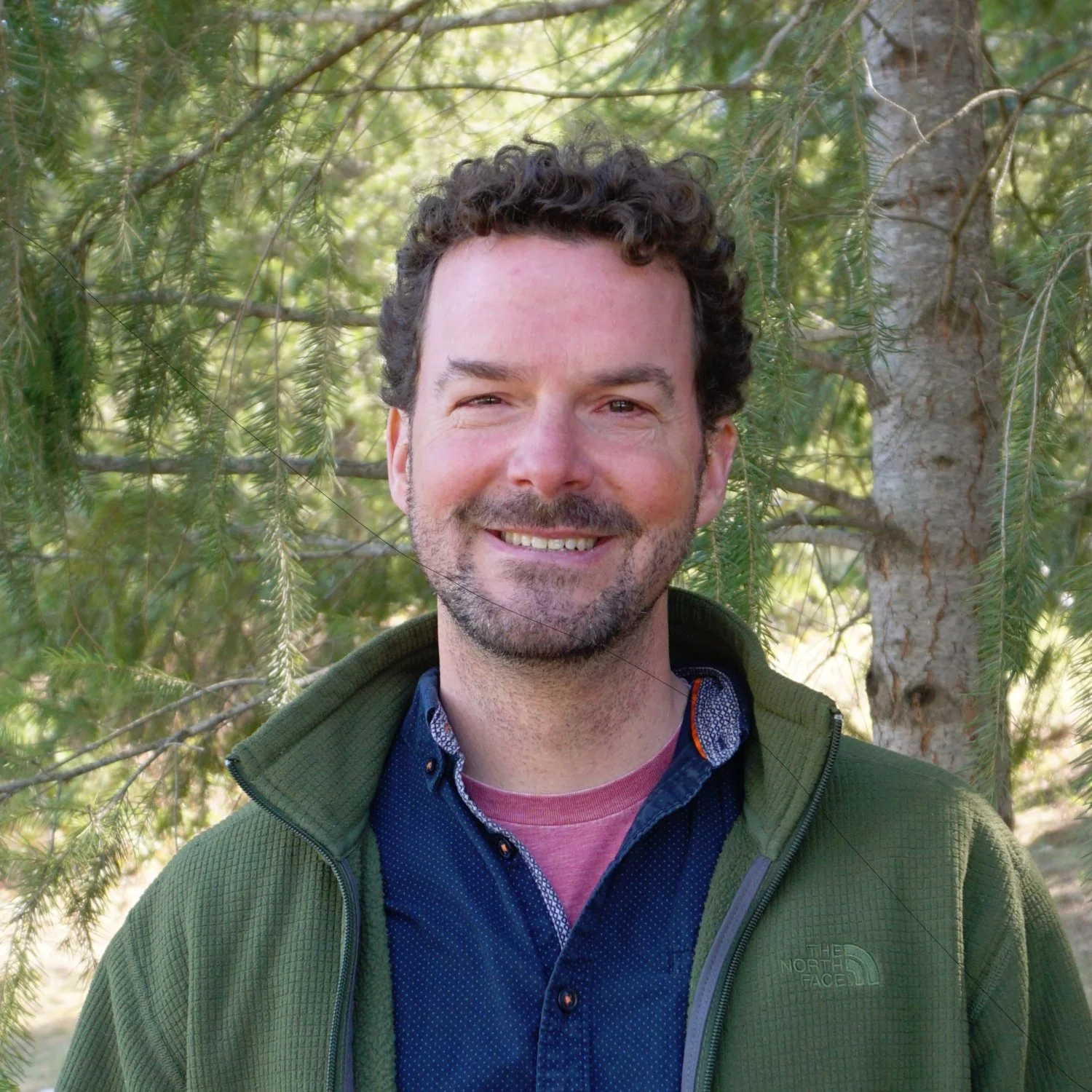 A smiling man with dark curly hair and a beard, wearing a green jacket and a blue shirt, standing in front of a pine tree with green branches and a tree trunk visible.