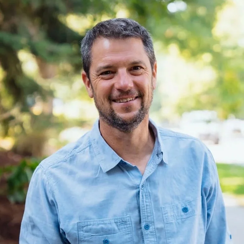 A smiling man with dark hair and a beard, wearing a light blue button-up shirt, standing outdoors with greenery in the background.
