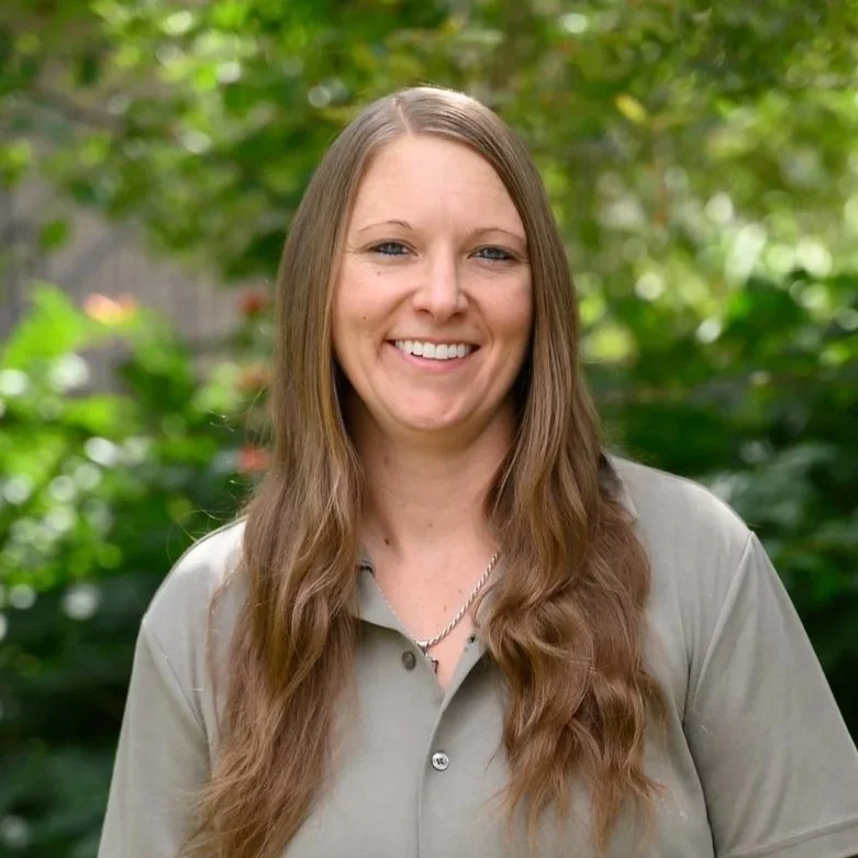 A smiling woman with long, wavy brown hair, wearing a grey collared shirt, standing outdoors with green foliage in the background.