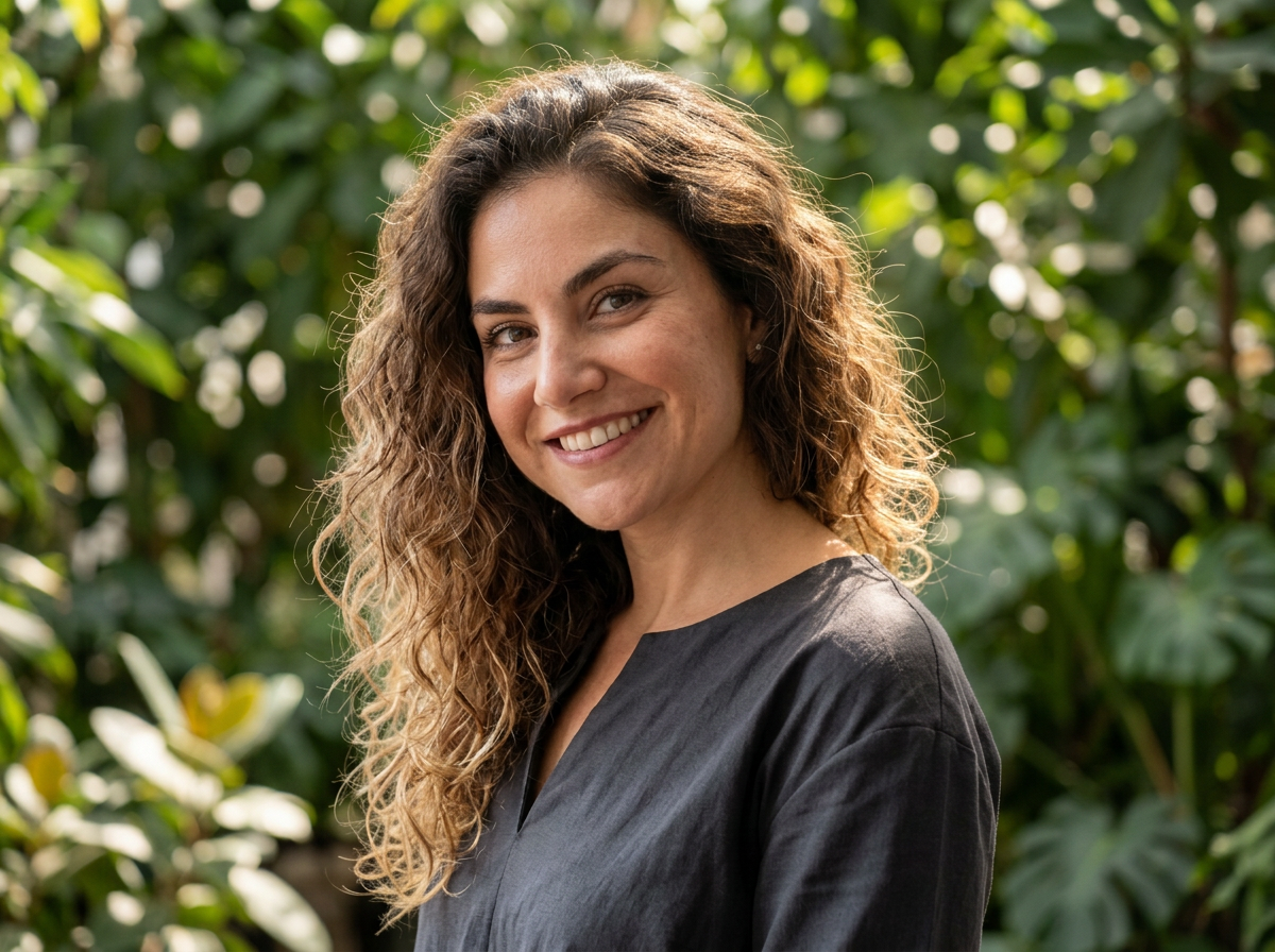 A woman with curly brown hair smiling outdoors in front of green foliage, wearing a dark-colored top.