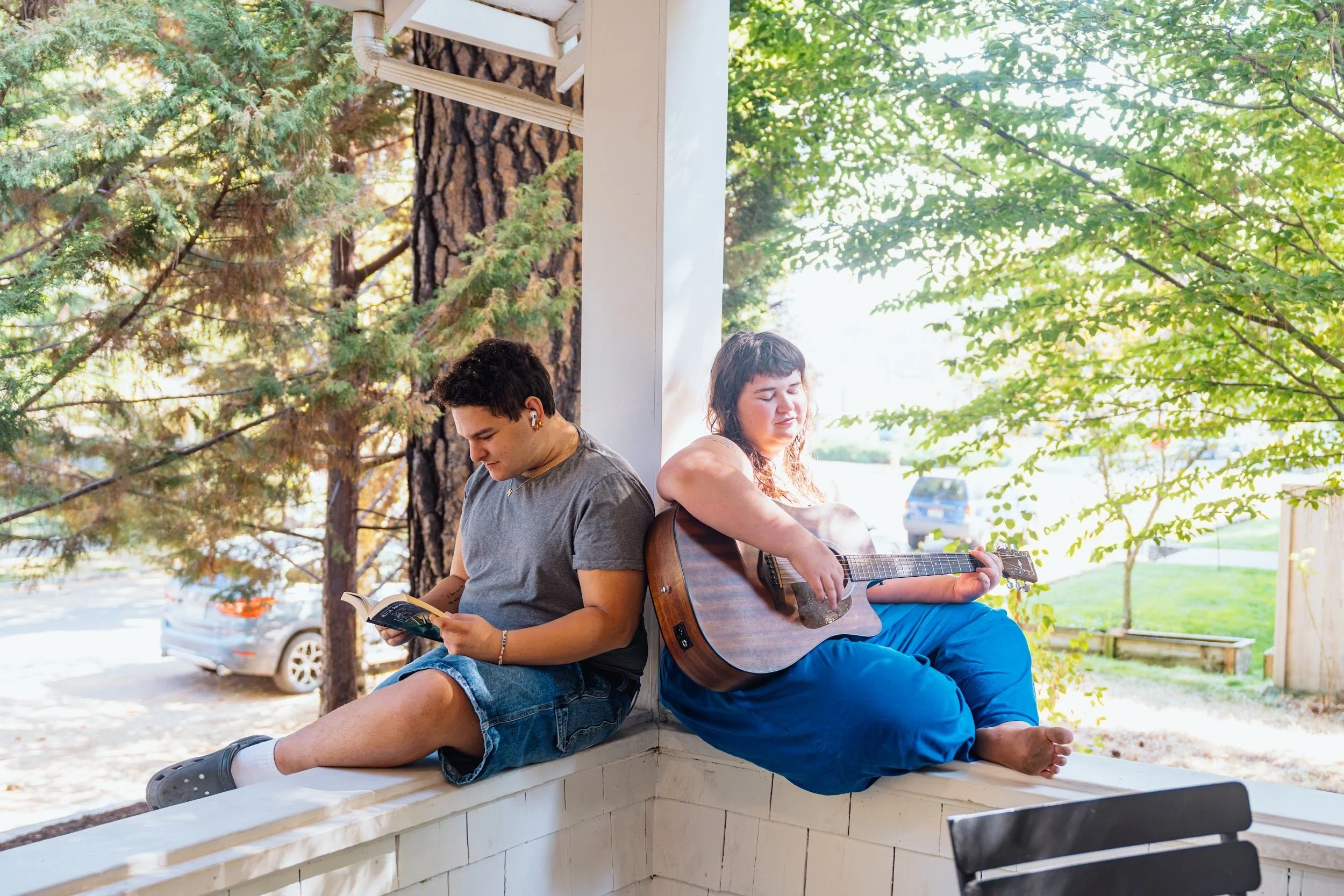 Two young people sitting back to back on a white porch, one reading a book and the other playing an acoustic guitar, with trees and parked cars in the background.