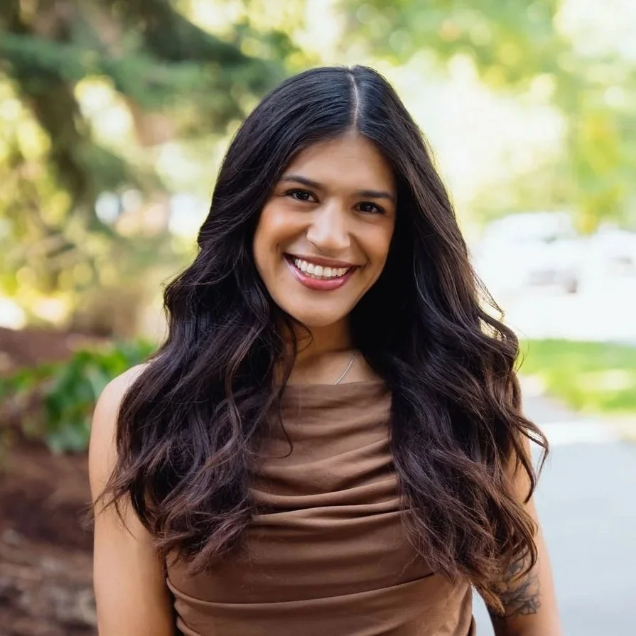 A woman smiling outdoors in a park with trees in the background, wearing a brown top.