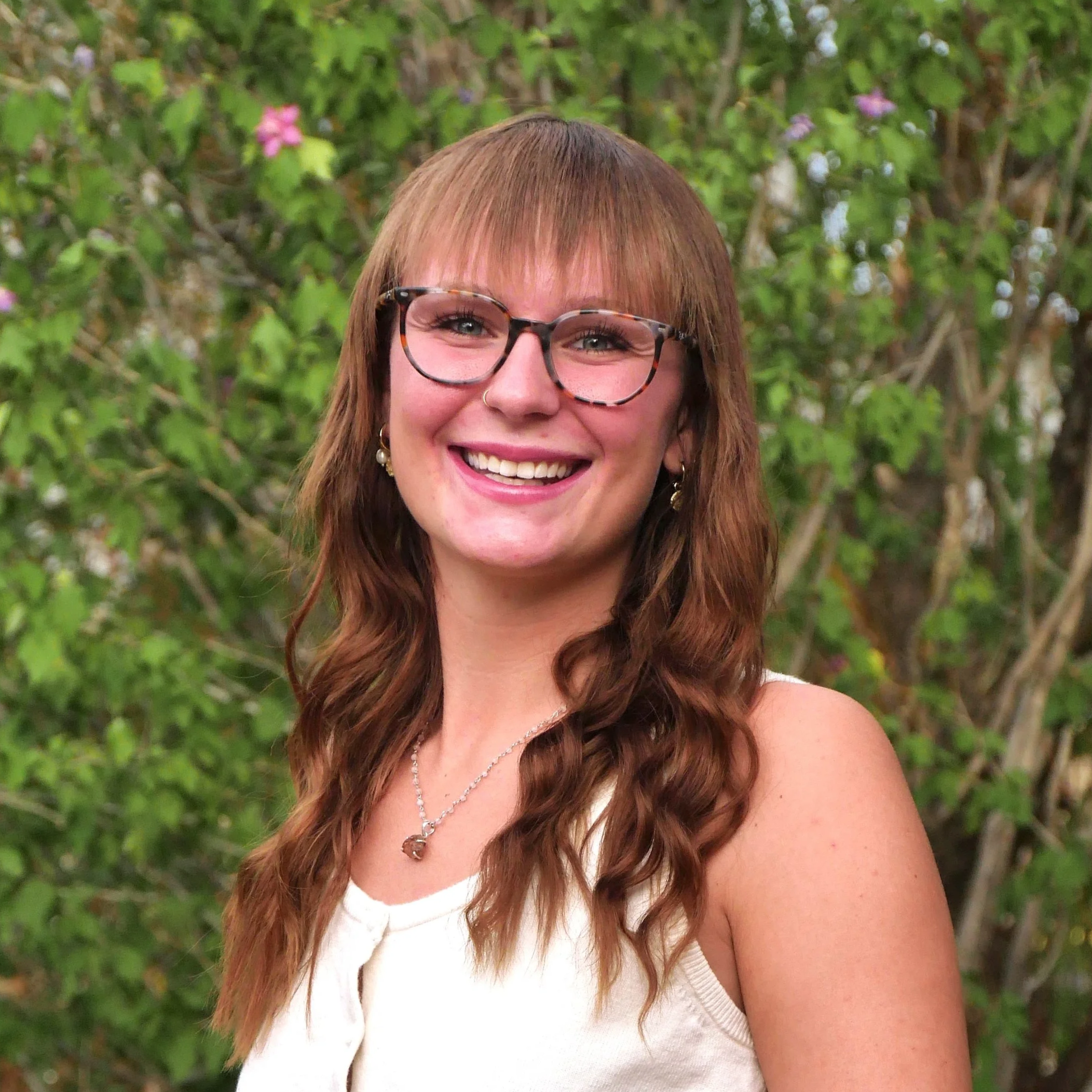Close-up portrait of a smiling woman with brown wavy hair, wearing glasses and earrings, outdoors with green foliage and pink flowers in the background.
