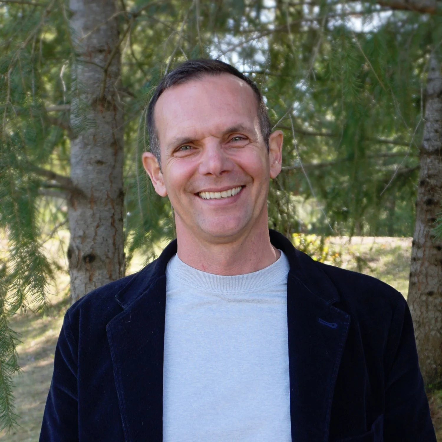 A smiling man with short dark hair and blue eyes standing outdoors in front of trees and greenery.