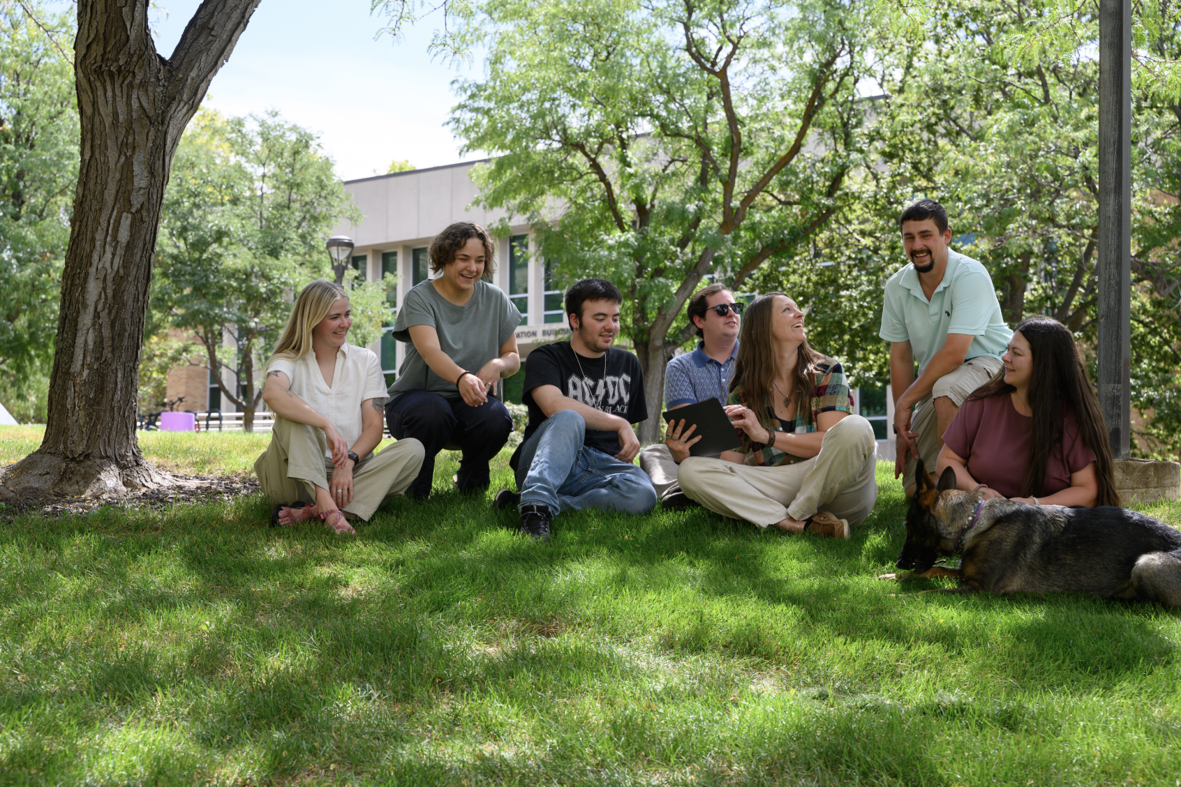 A group of eight young people sitting and kneeling on grass under a large tree, sharing a moment of laughter and conversation outdoors on a sunny day, with a dog lying nearby.