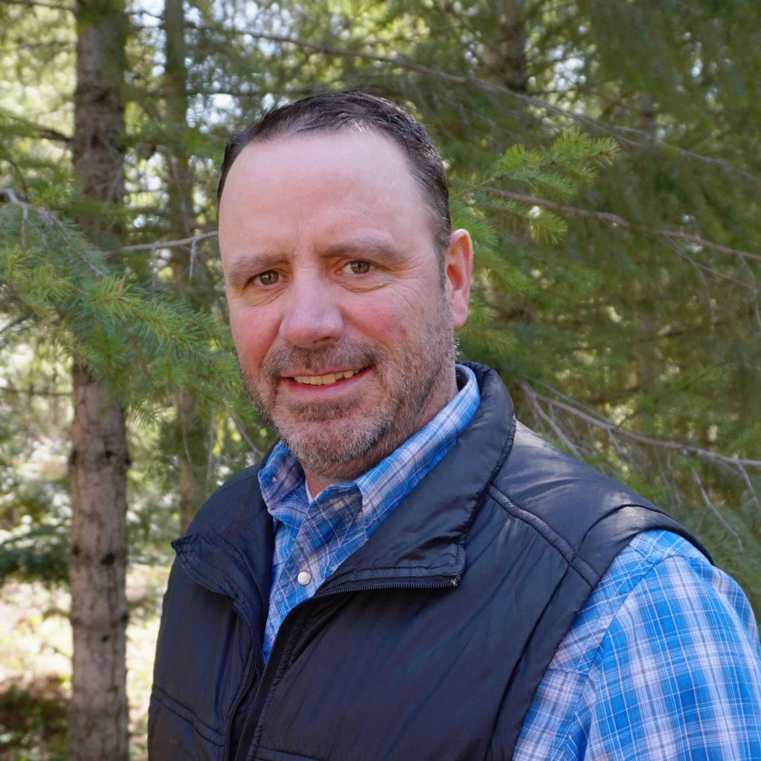 A man with short dark hair and a beard smiling outdoors in front of green trees, wearing a blue plaid shirt and a black vest.