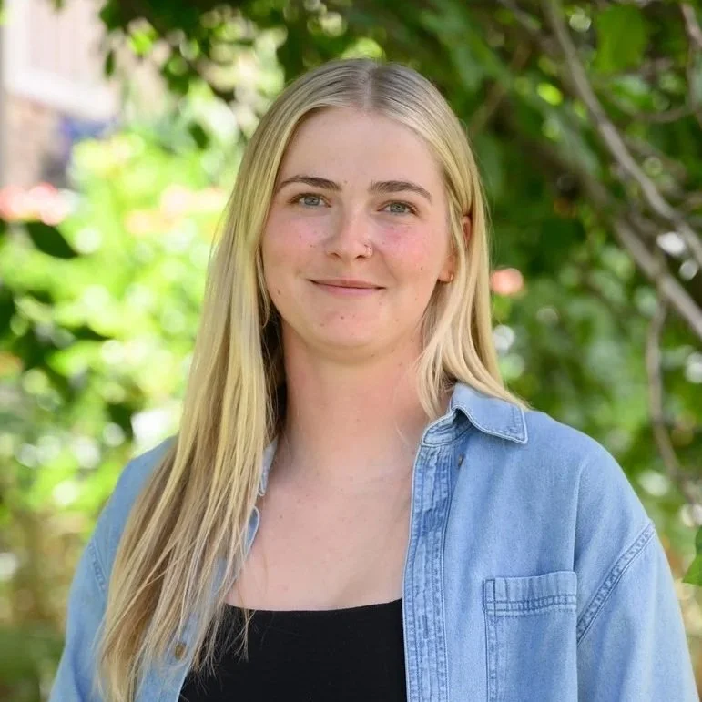 A young woman with long blonde hair and a nose piercing, smiling outdoors with green foliage in the background, wearing a black top and denim shirt.